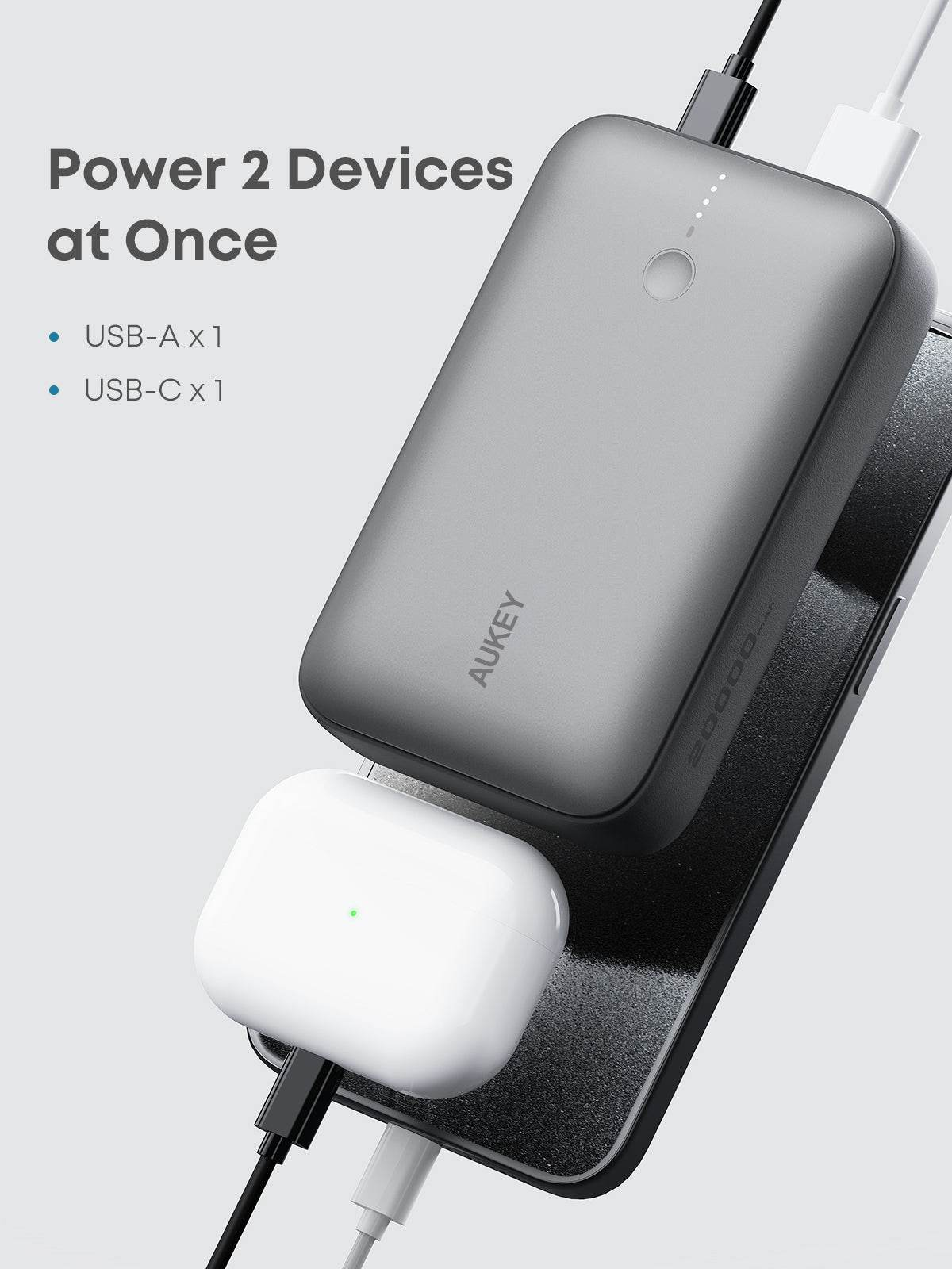 Un power bank grigio carica un telefono e auricolari bianchi. Testo: 'Power 2 Devices at Once'. Logo AUKEY visibile.