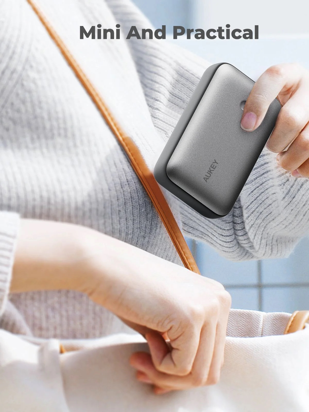Una persona tiene in mano un power bank grigio, vicino a un cinturino di borsa marrone chiaro. Il power bank ha il marchio 'AUKEY'. Lo sfondo è un maglione grigio.