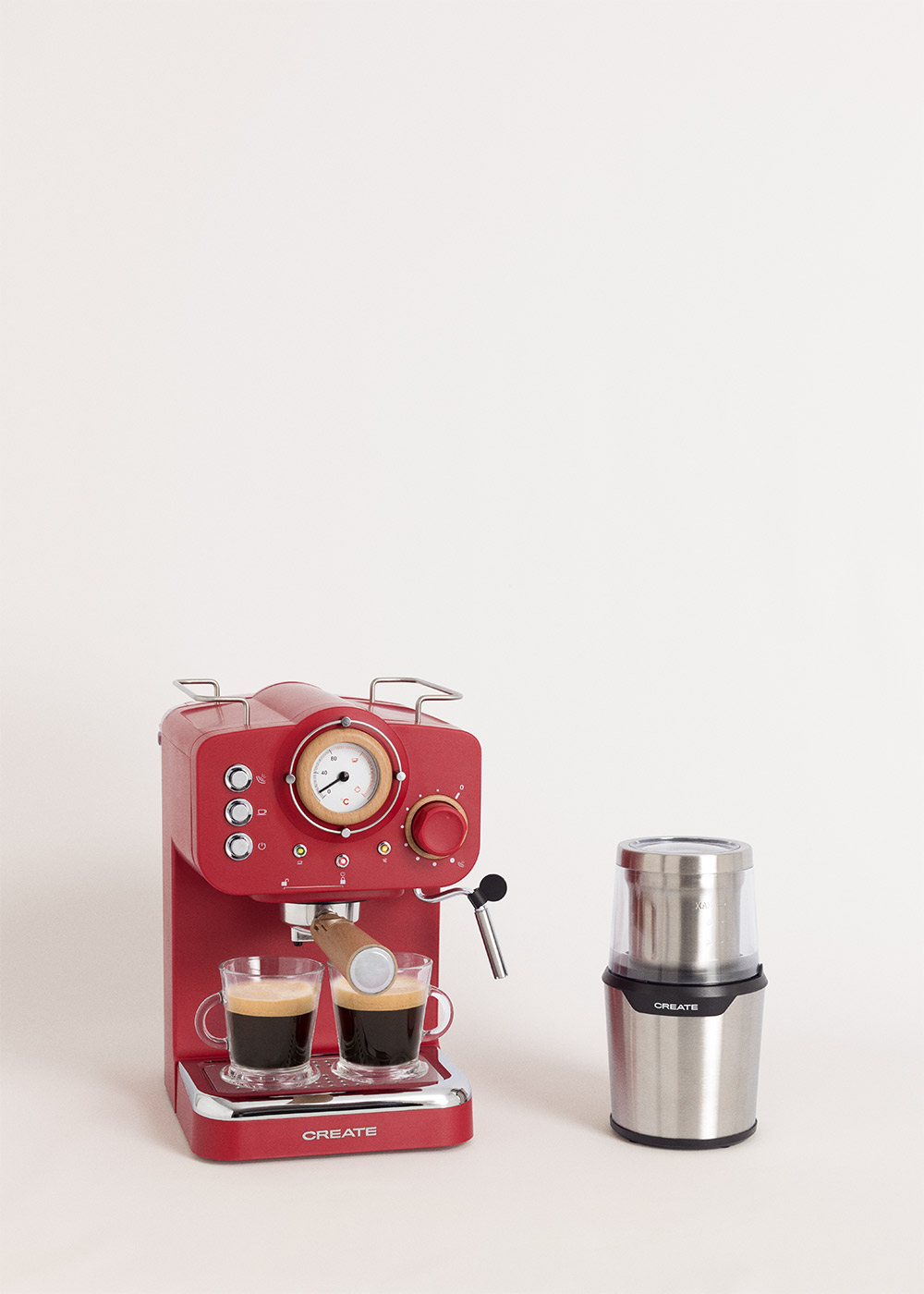 Una cafetera espresso roja con dos tazas de café junto a un molinillo de café plateado sobre fondo blanco.