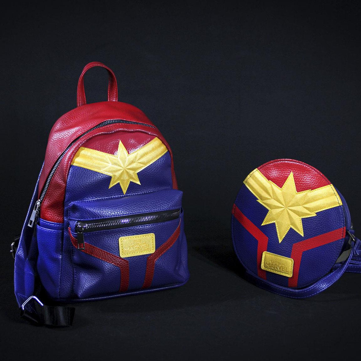 Dos bolsos de Capitana Marvel sobre fondo negro. Uno es una mochila, el otro un bolso redondo, ambos azules, rojos y dorados.
