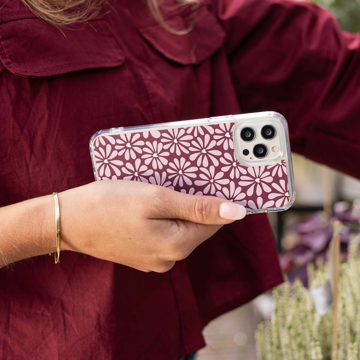 Een vrouw houdt een telefoon vast met een bloemenhoesje. Ze draagt een bordeauxrode blouse en een gouden armband. Planten op de achtergrond.