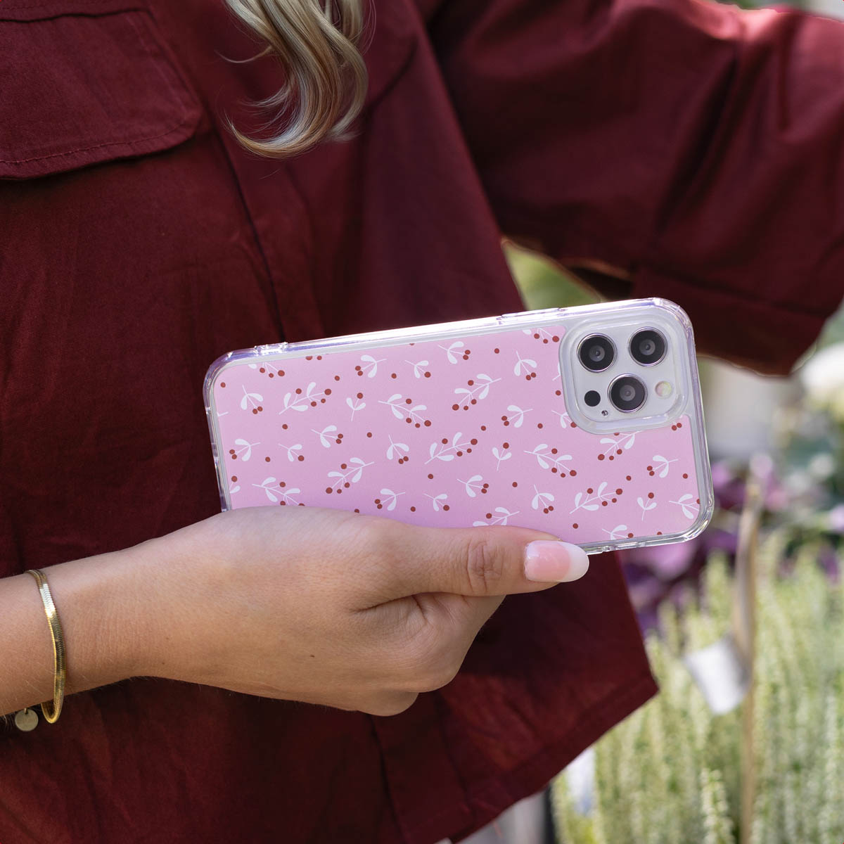 Een persoon houdt een telefoon vast met een roze bloemenhoesje. De persoon draagt een bordeauxrode blouse en een gouden armband. Op de achtergrond zijn planten te zien.