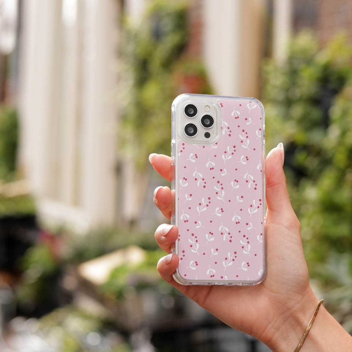 Een hand houdt een telefoon met een bloemenhoes vast. De hoes is roze met witte bladeren en rode bessen. De achtergrond is wazig groen.