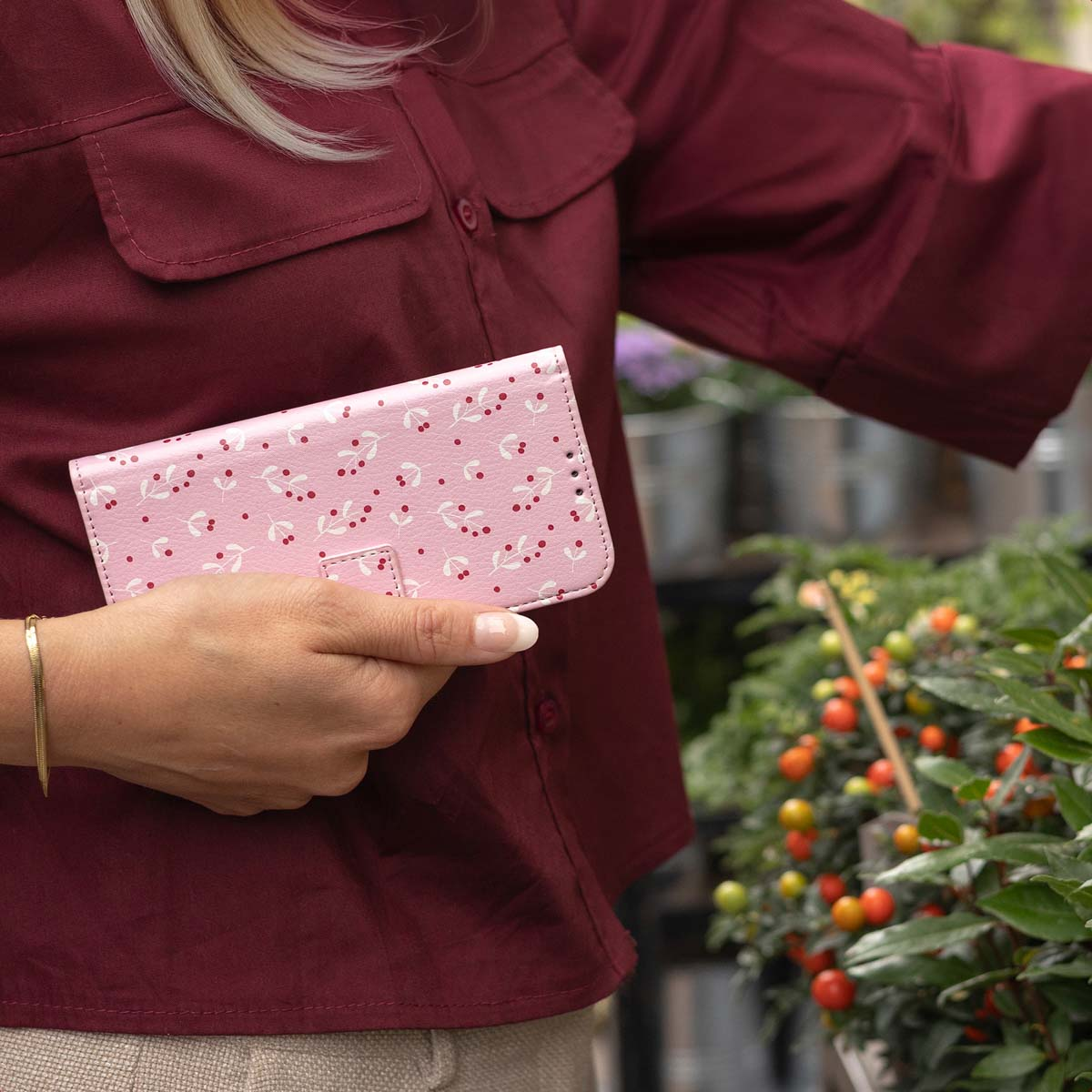 Vrouw houdt roze telefoonhoes vast met bessenpatroon. Ze draagt een bordeauxrode blouse en staat bij planten met rode en oranje bessen.