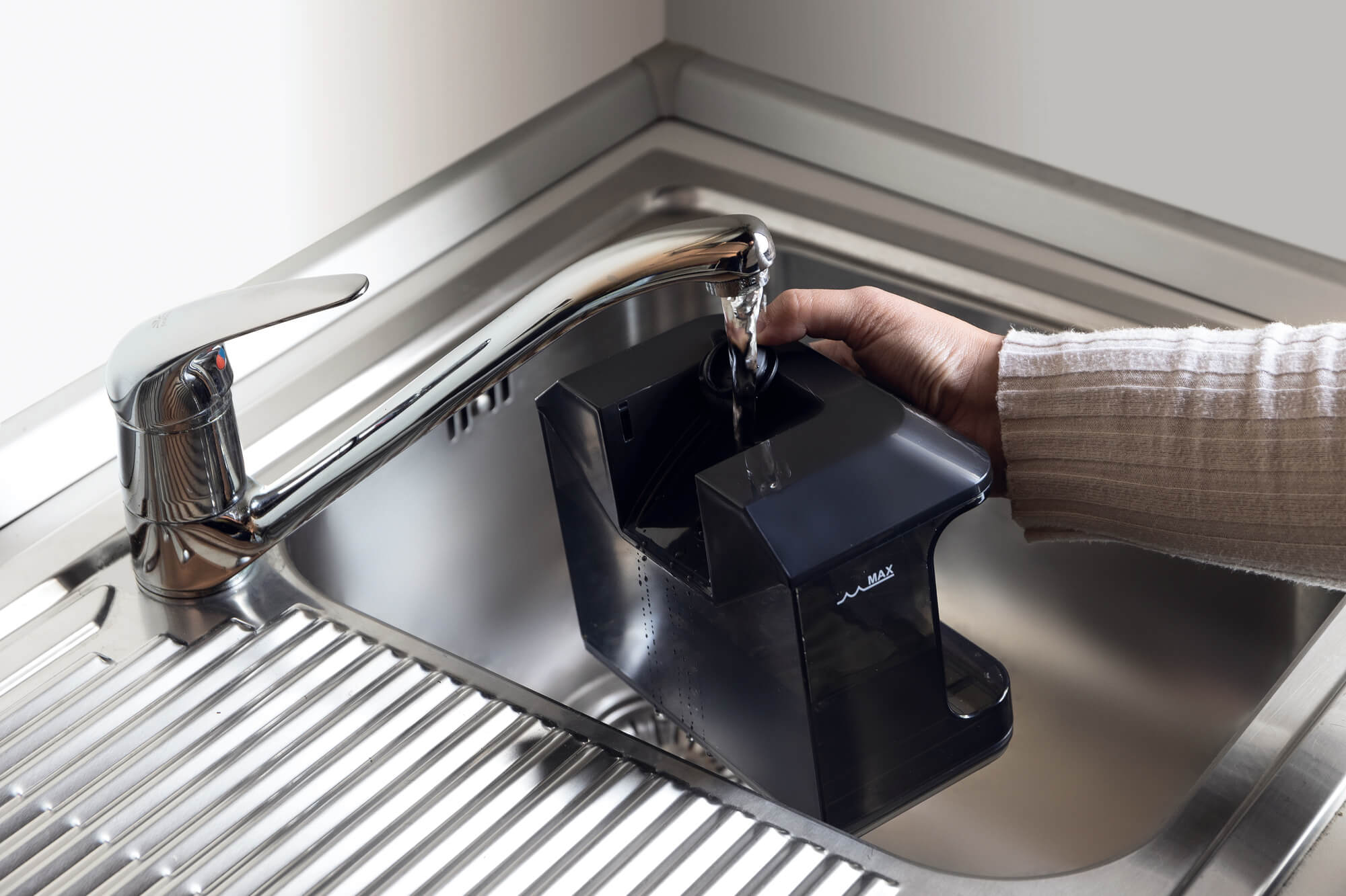 Eine Hand füllt einen schwarzen Behälter mit Wasser aus einem verchromten Wasserhahn in einem Edelstahlbecken. Der Behälter sagt MAX.