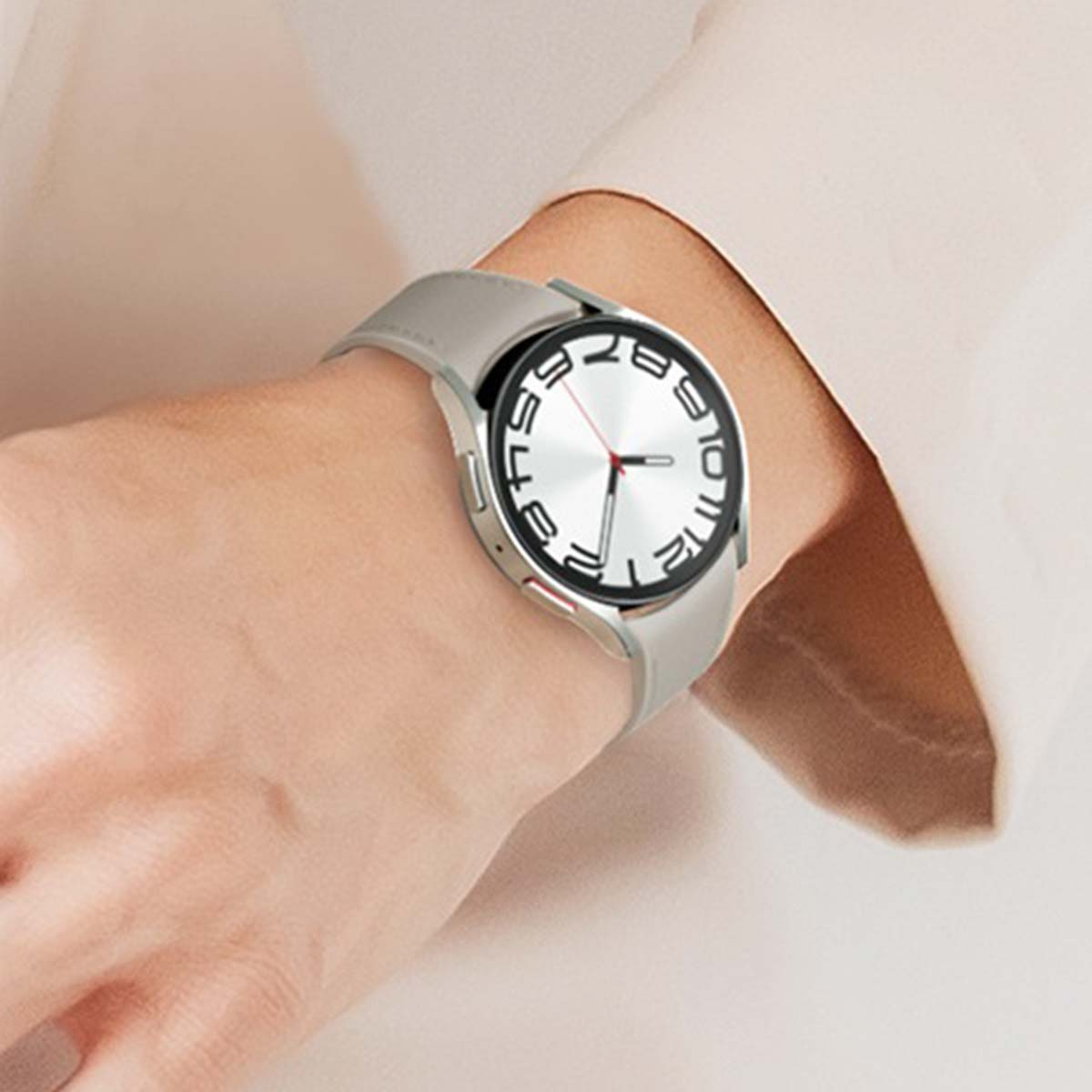 Nadgarstek osoby ze srebrnym smartwatchem. Zegarek ma białą tarczę z czarnymi cyframi i szary pasek. Tło jest beżowe.