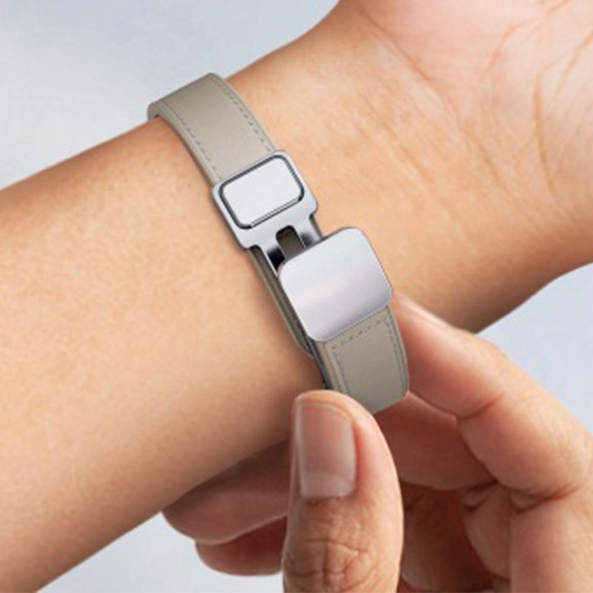 Nadgarstek osoby z beżowym skórzanym zegarkiem. Zegarek ma srebrne zapięcie. Druga ręka trzyma zegarek.