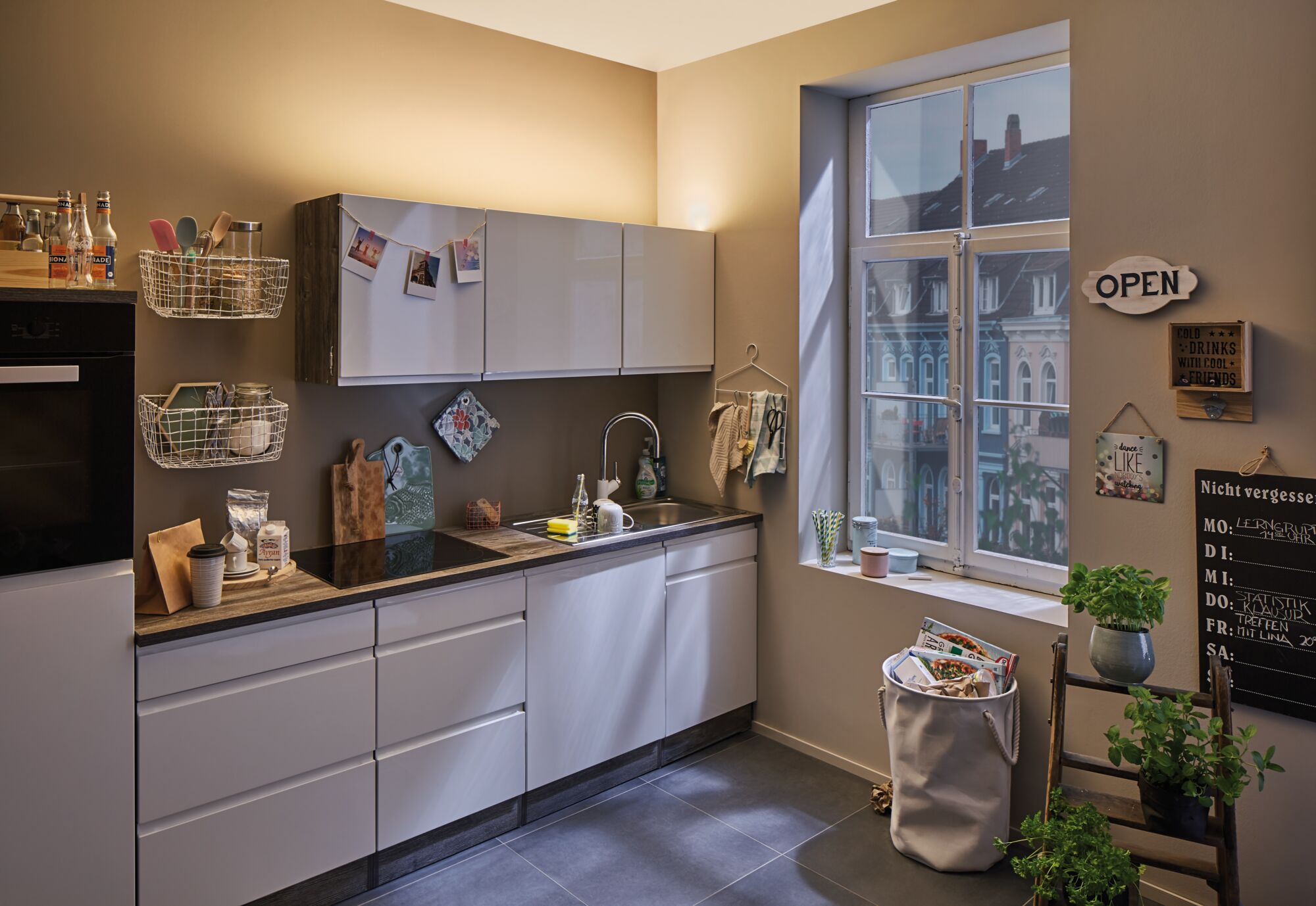 Cuisine avec des armoires blanches, un évier, une fenêtre avec un paysage urbain et diverses décorations. Il y a un four noir et une échelle avec des plantes.