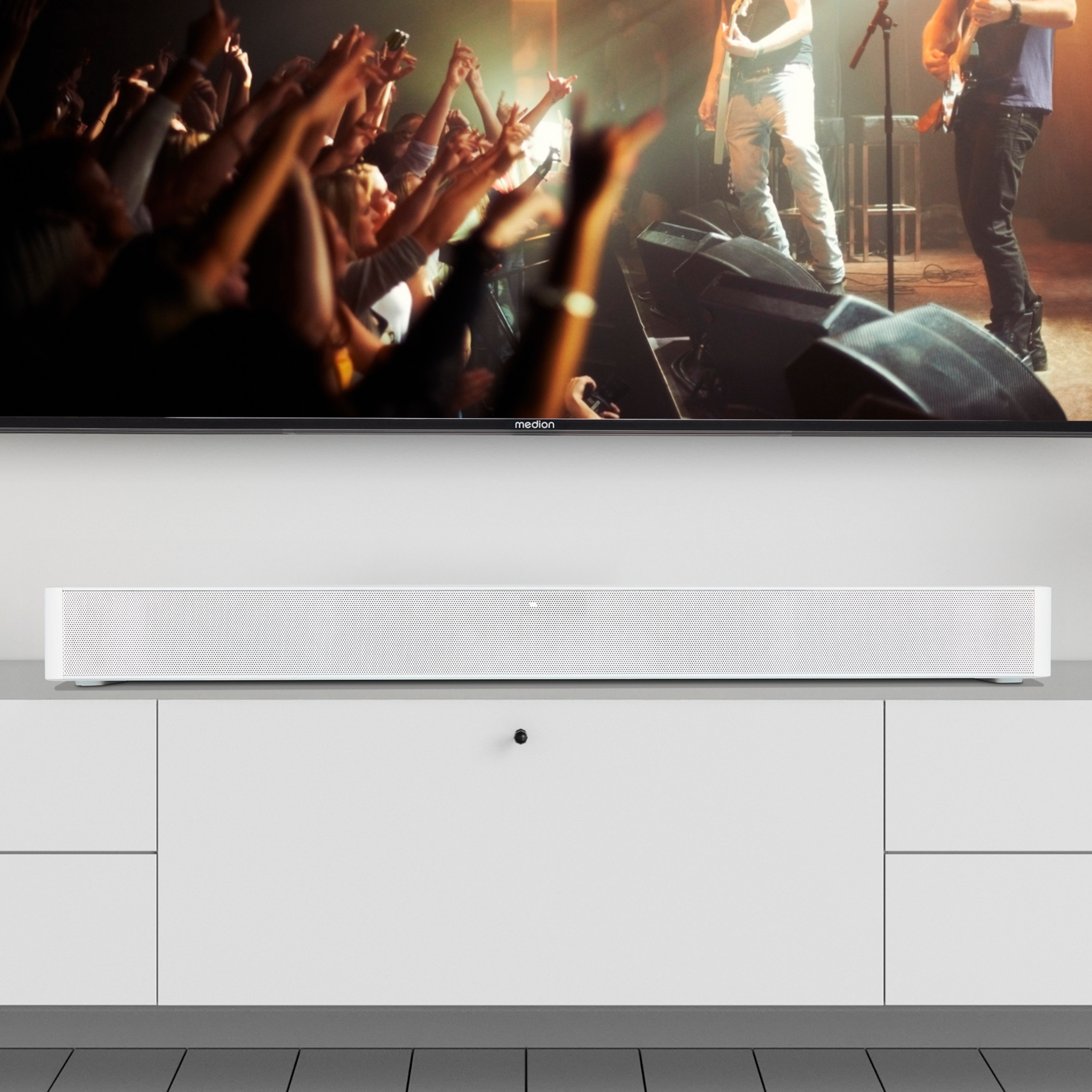 Ein Fernseher zeigt ein Konzert mit erhobenen Händen des Publikums. Eine weiße Soundbar steht auf einem weißen Schrank.
