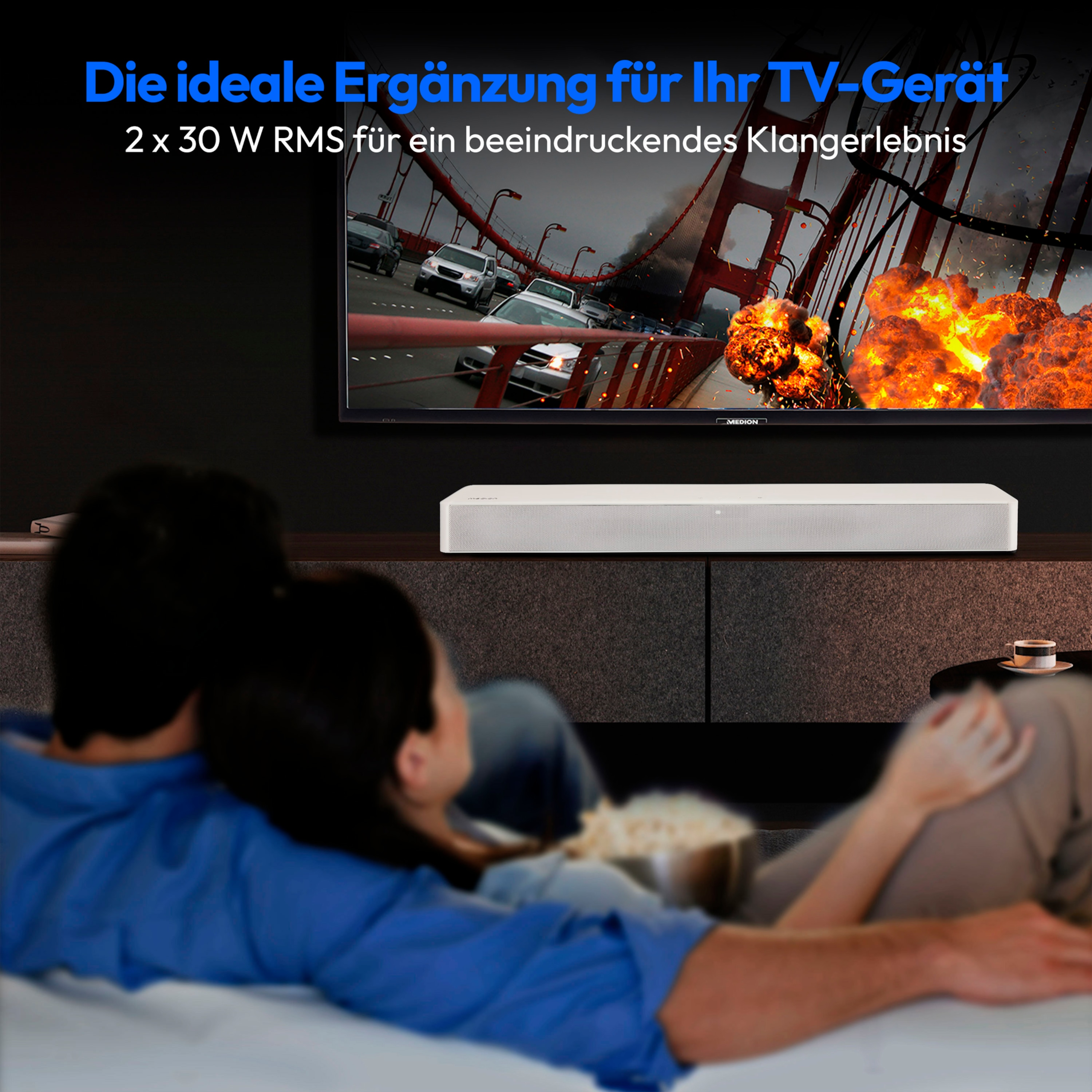 Ein Paar sieht fern. Eine Soundbar steht darunter. Der Bildschirm zeigt eine Katastrophenszene.