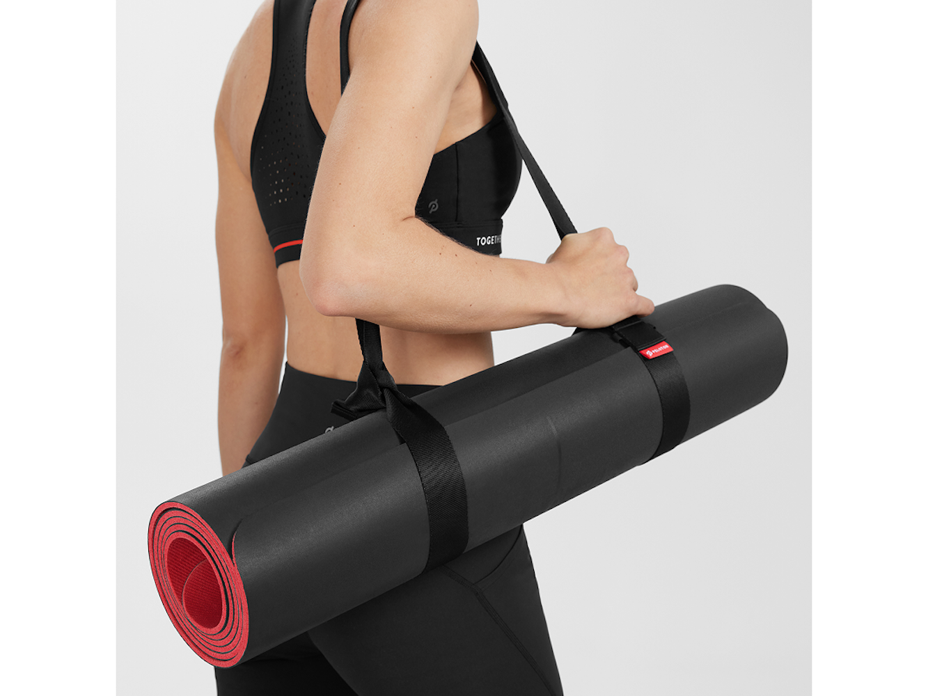 Eine Frau in Sportkleidung trägt eine schwarze Yogamatte mit roter Innenseite. Die Matte ist umgeschnallt und über ihrer Schulter gehalten.