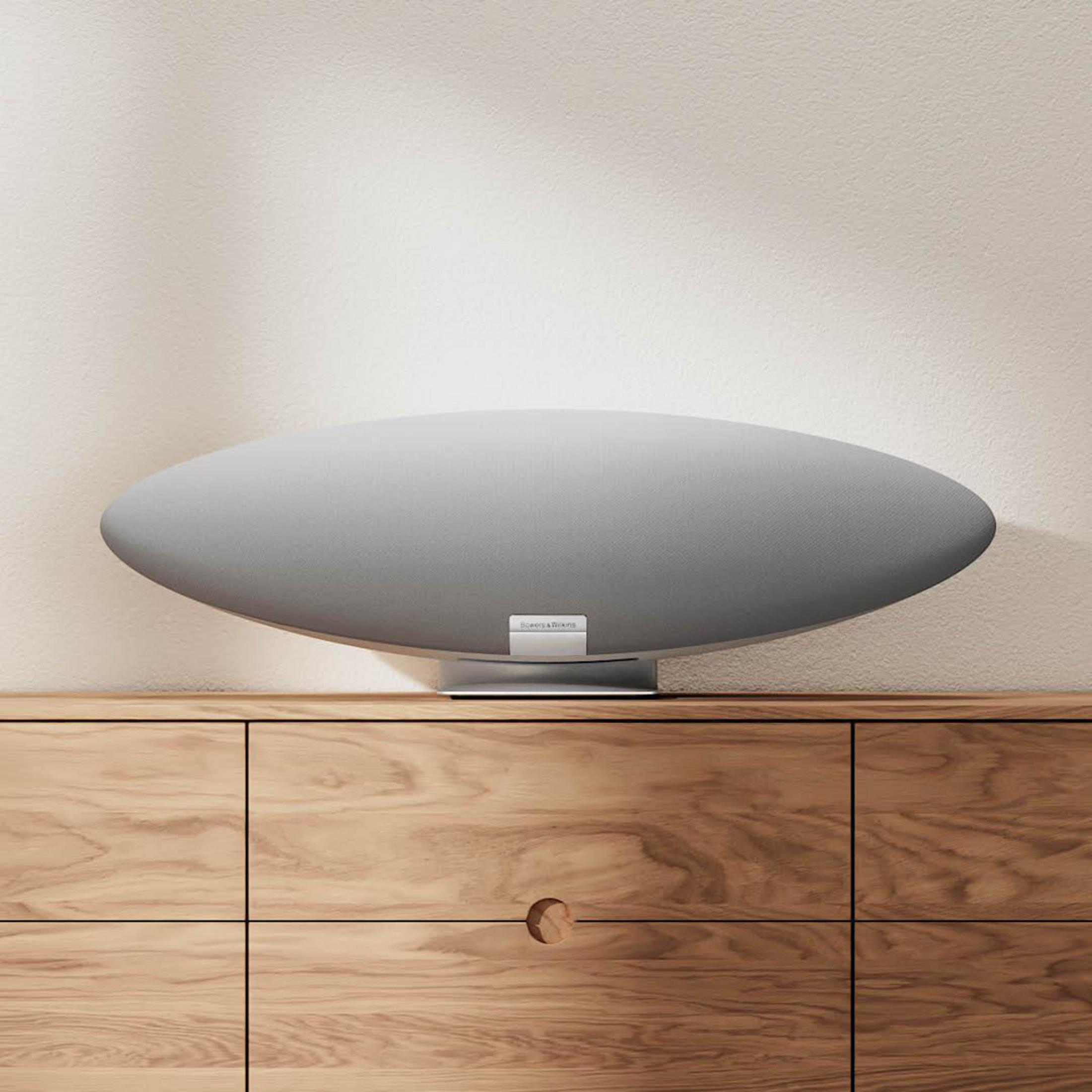 Ovale graue Lautsprecher auf Holzschrank vor strukturierter beiger Wand. Der Lautsprecher hat einen Metallsockel.