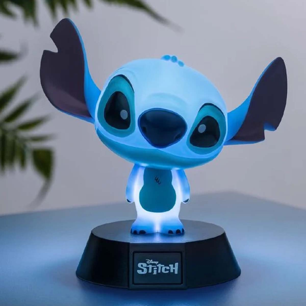 Eine blaue Stitch-Lampe steht auf einem schwarzen Sockel. Sie hat große Ohren, große Augen und einen leuchtenden Körper. Die Lampe ist beleuchtet.