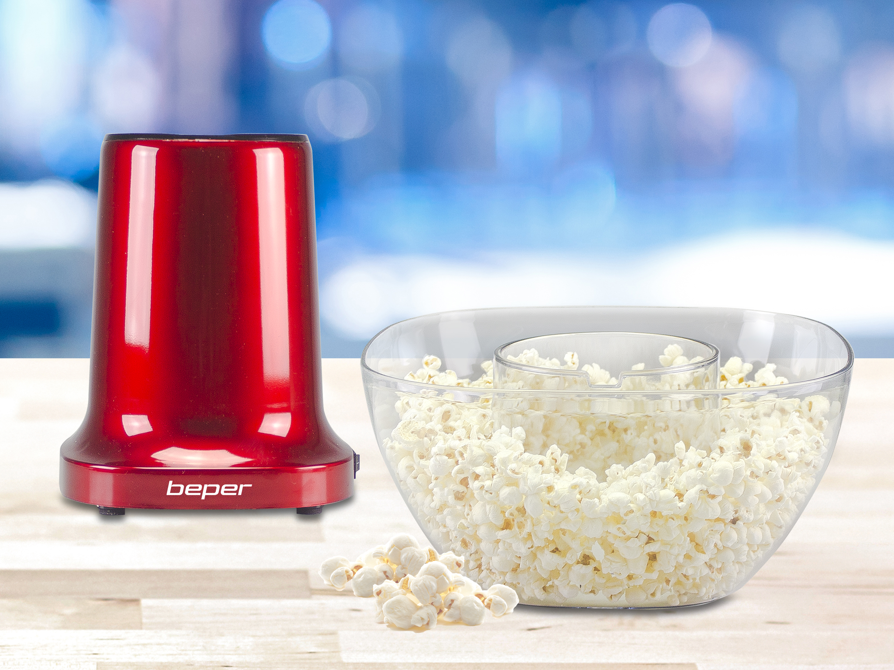 Una macchina per popcorn rossa e una ciotola di popcorn sono su un tavolo. Lo sfondo è sfocato blu e bianco.