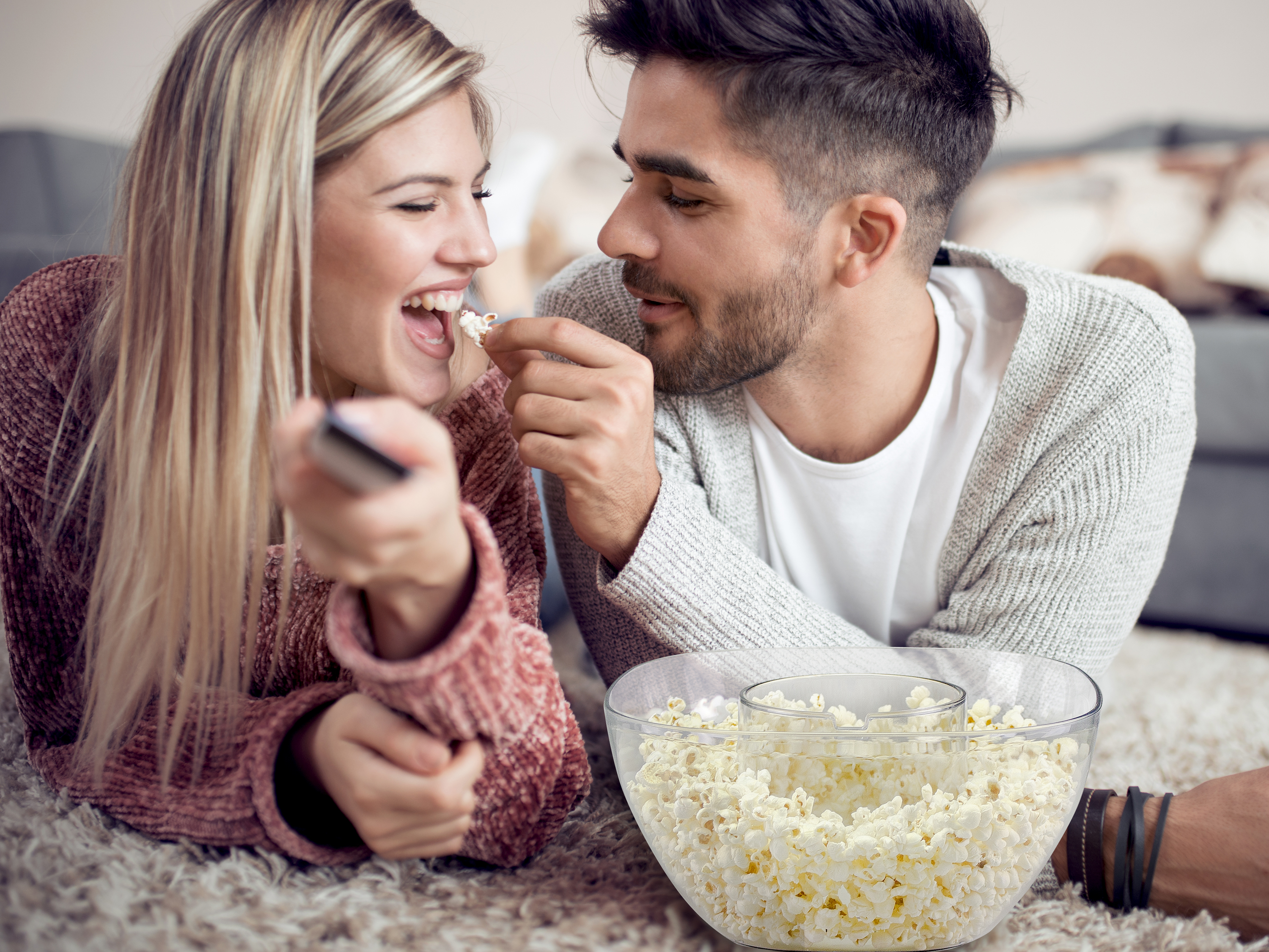 Una coppia sul pavimento mangia popcorn. Donna con telecomando, uomo che la nutre. Ciotola di popcorn davanti.