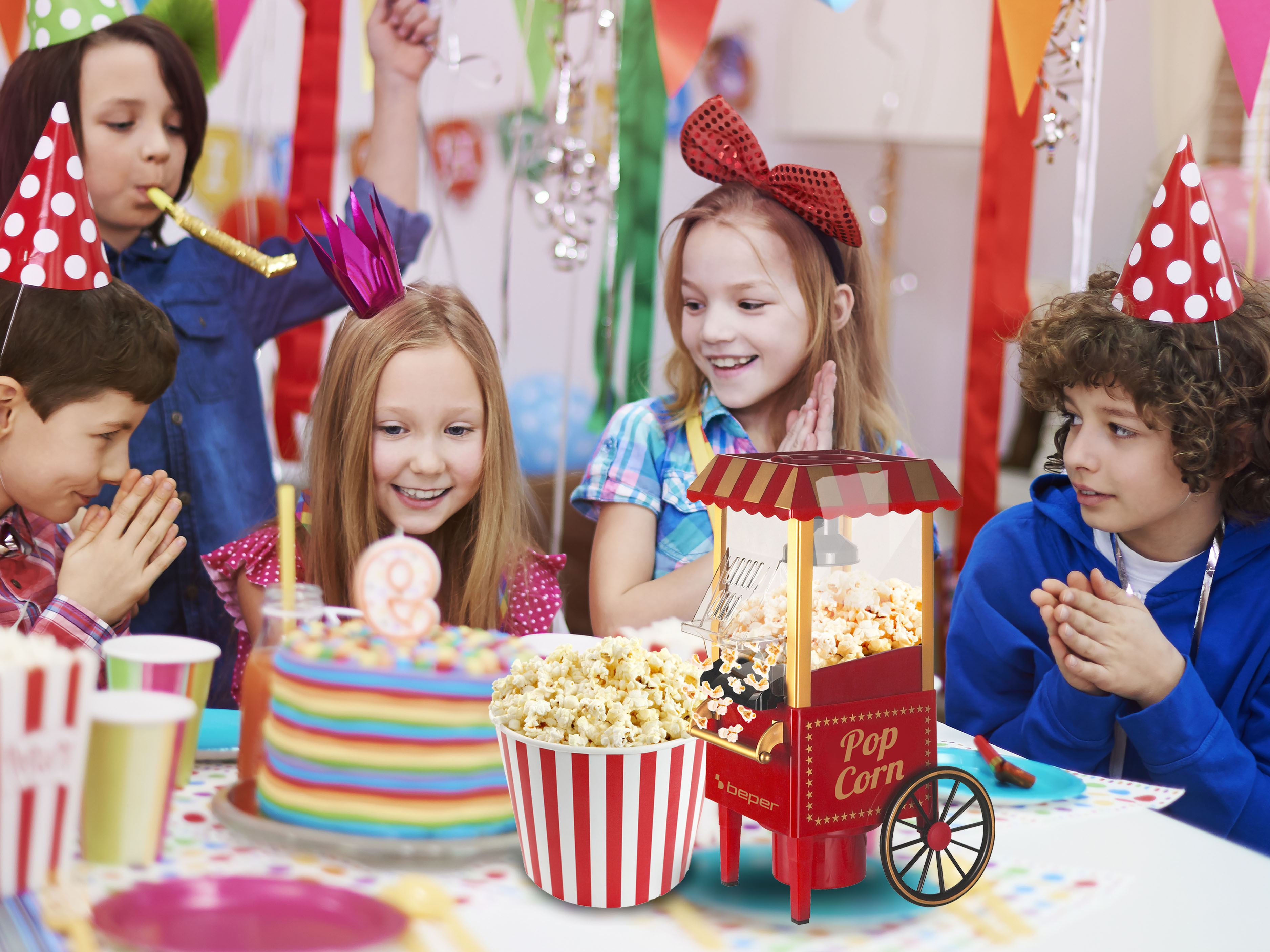 Bambini a una festa con torta, popcorn e cappellini. Alcuni applaudono. Una macchina per popcorn è al centro.