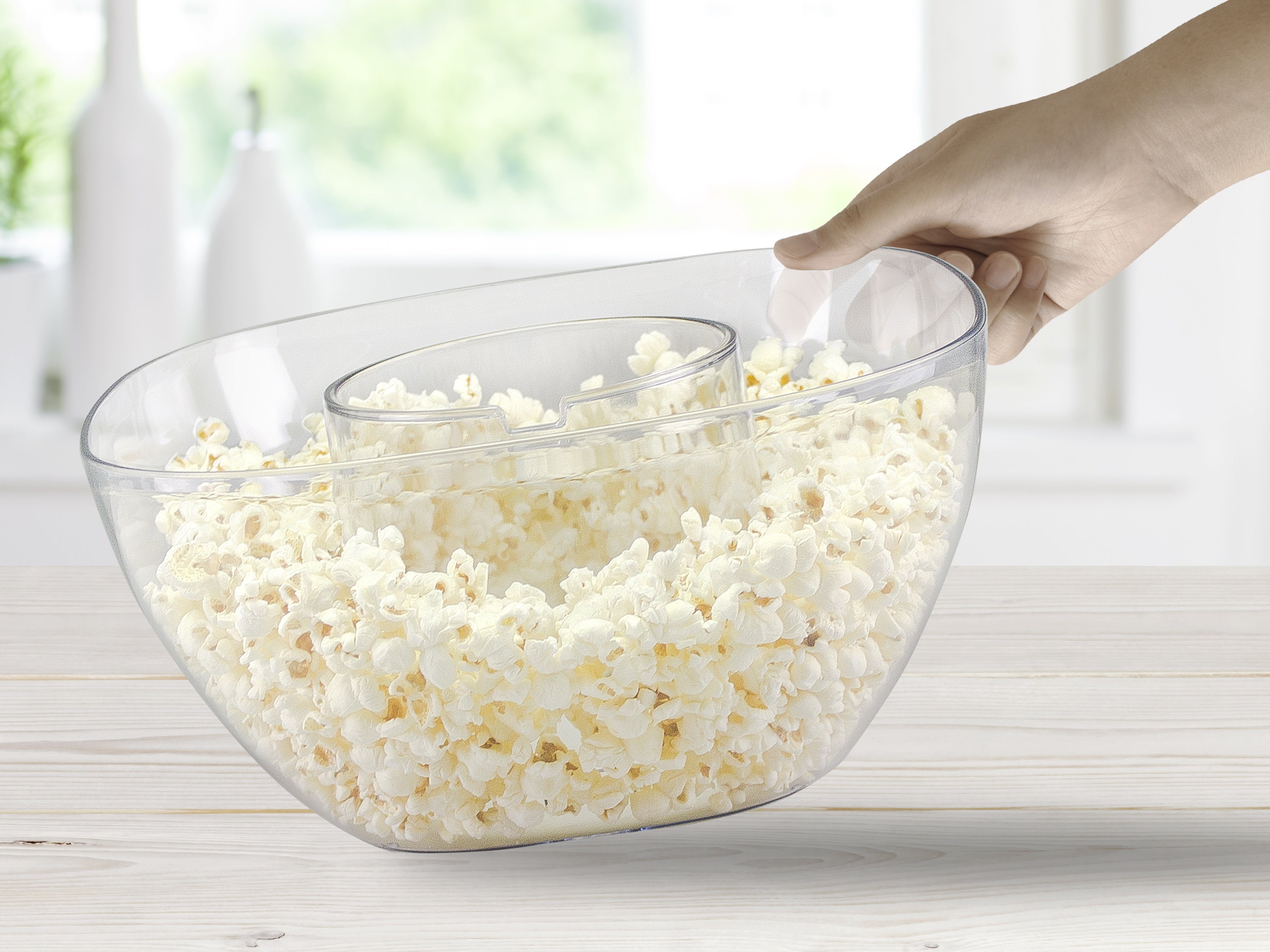 Una mano tiene una ciotola trasparente piena di popcorn con un vano centrale. Sfondo bianco con due vasi.