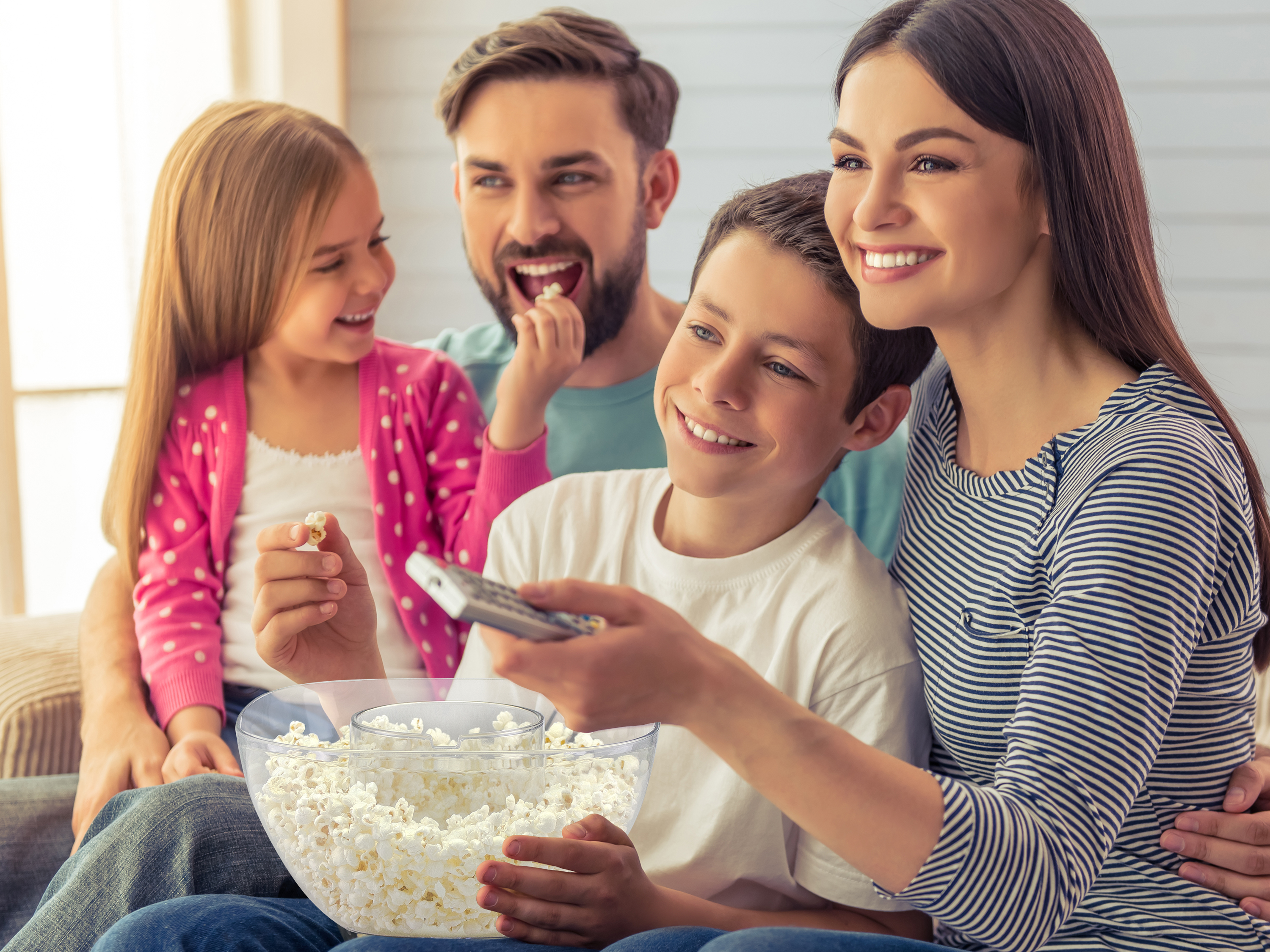 Famiglia guarda la TV, sorridendo. Figlio con telecomando, popcorn. Mamma abbraccia il figlio. Papà e figlia sorridono.