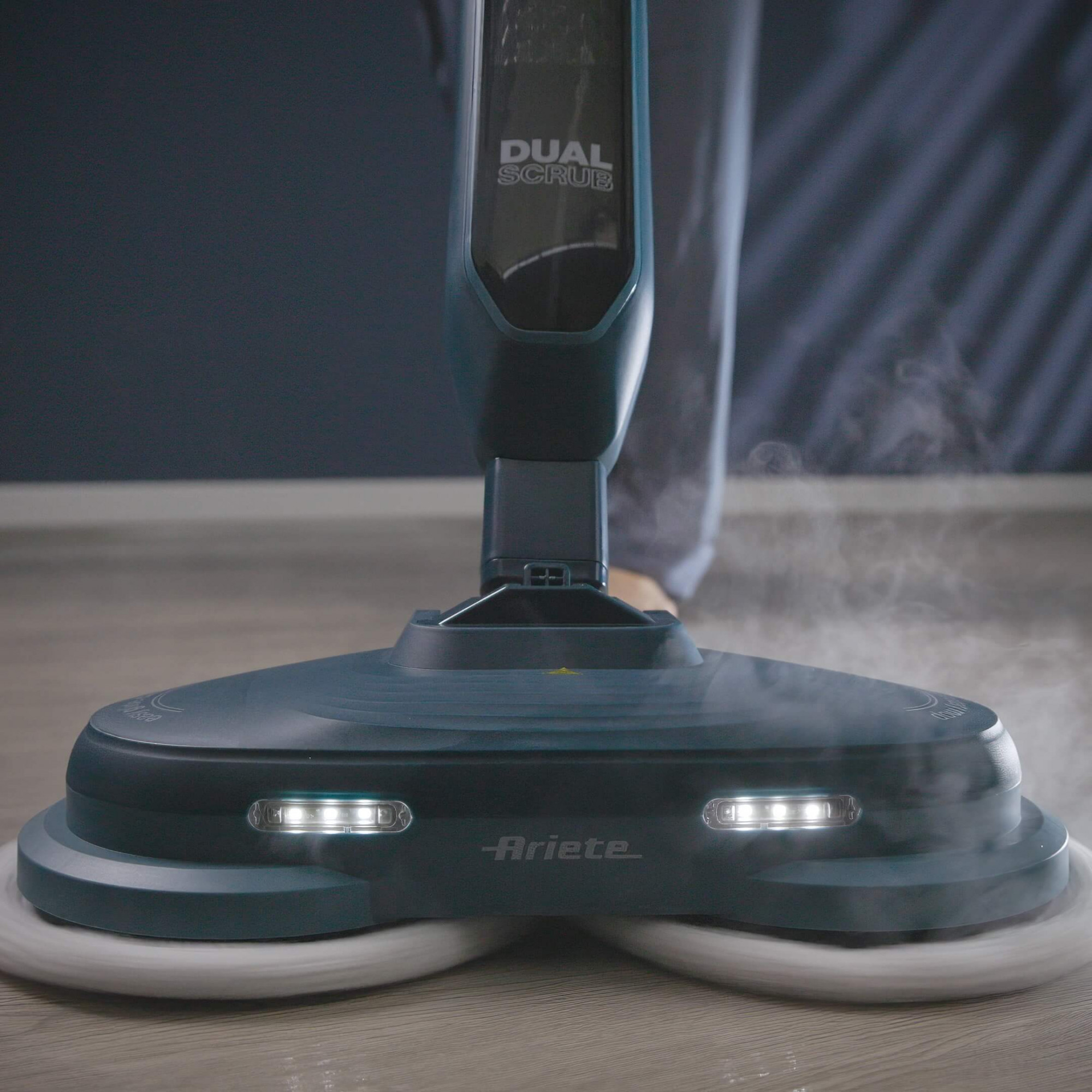 Ein türkisfarbener Dual Scrub Dampfmopp reinigt einen Holzboden. Dampf steigt auf. Der Mopp hat Lichter und das Ariete-Logo.