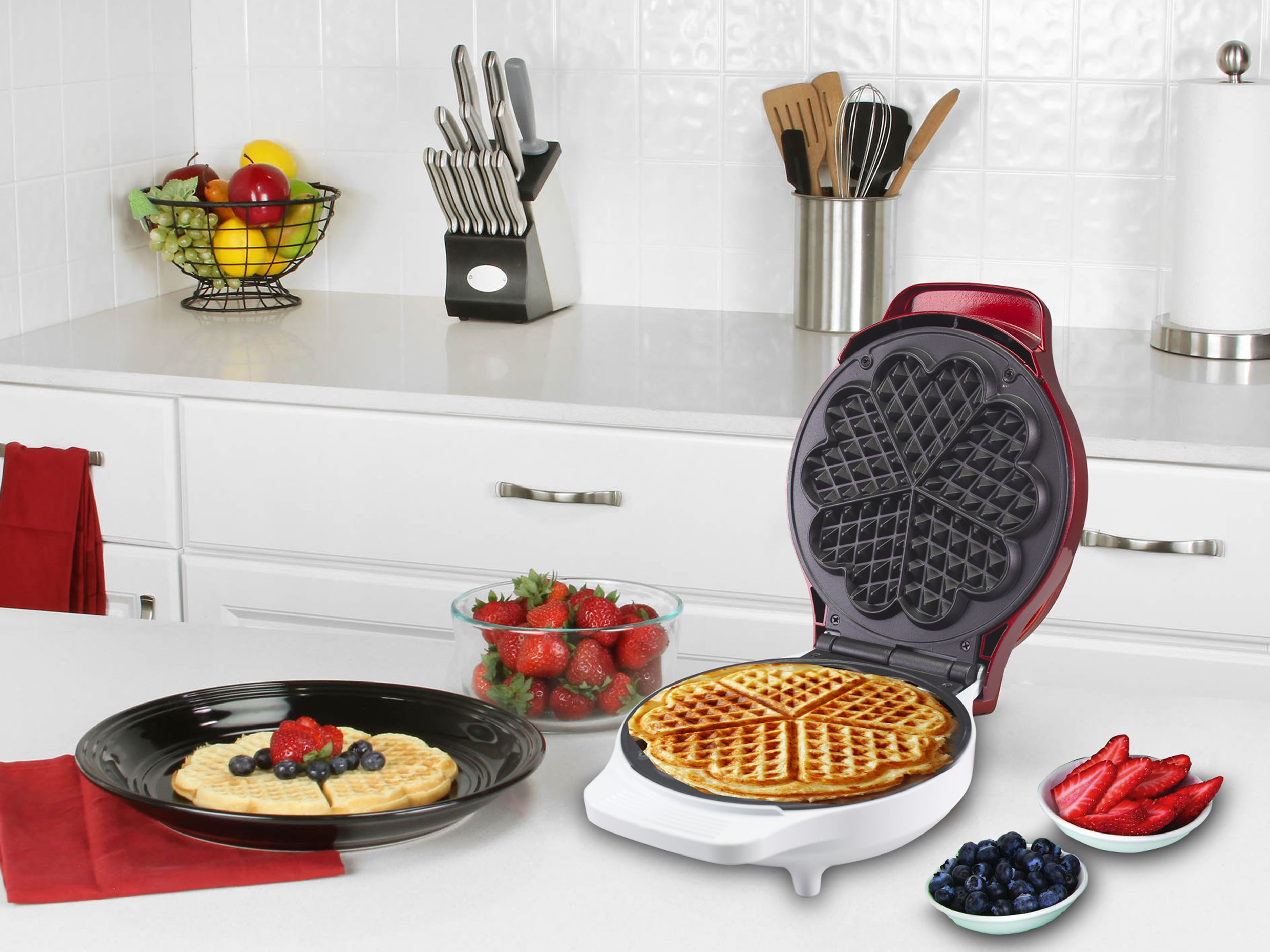 Una macchina per waffle rossa è aperta, accanto a un waffle su un piatto nero con fragole, mirtilli e frutta.