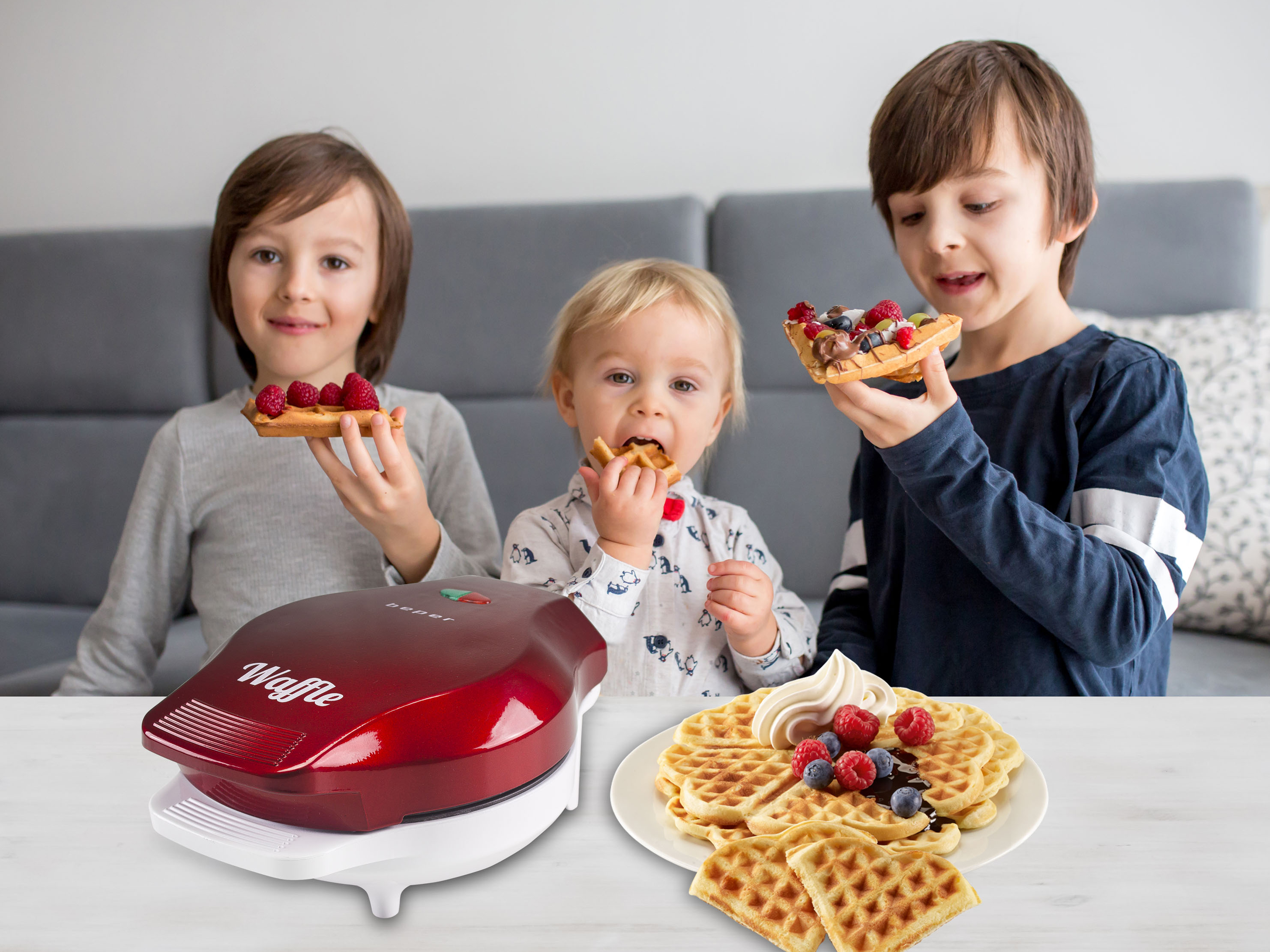 Tre bambini con waffle, uno mangia. I waffle hanno frutta. Una piastra per waffle rossa è su un tavolo bianco.
