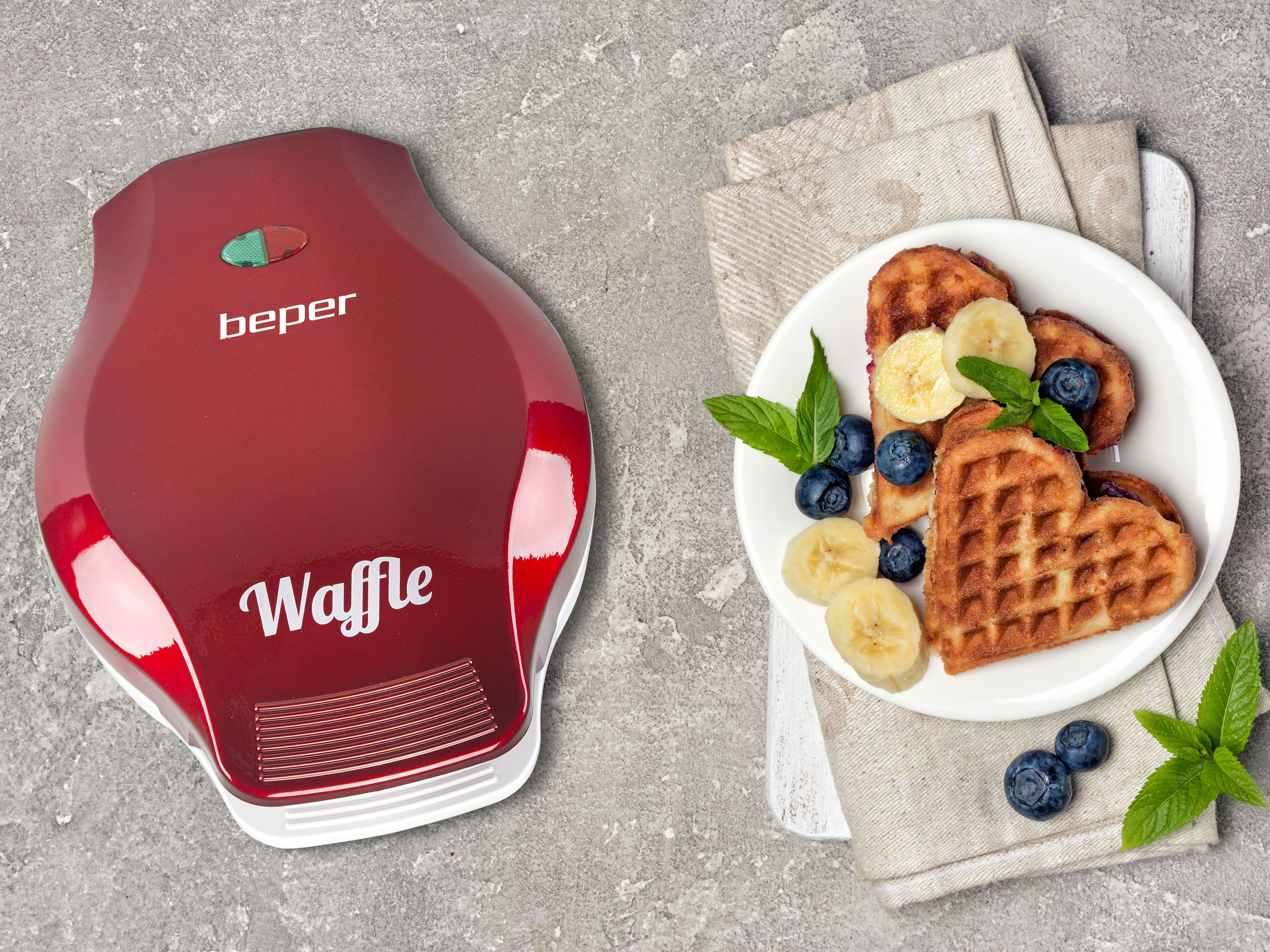 Una macchina per waffle rossa e waffle con mirtilli e banane su un piatto bianco, su una superficie grigia.