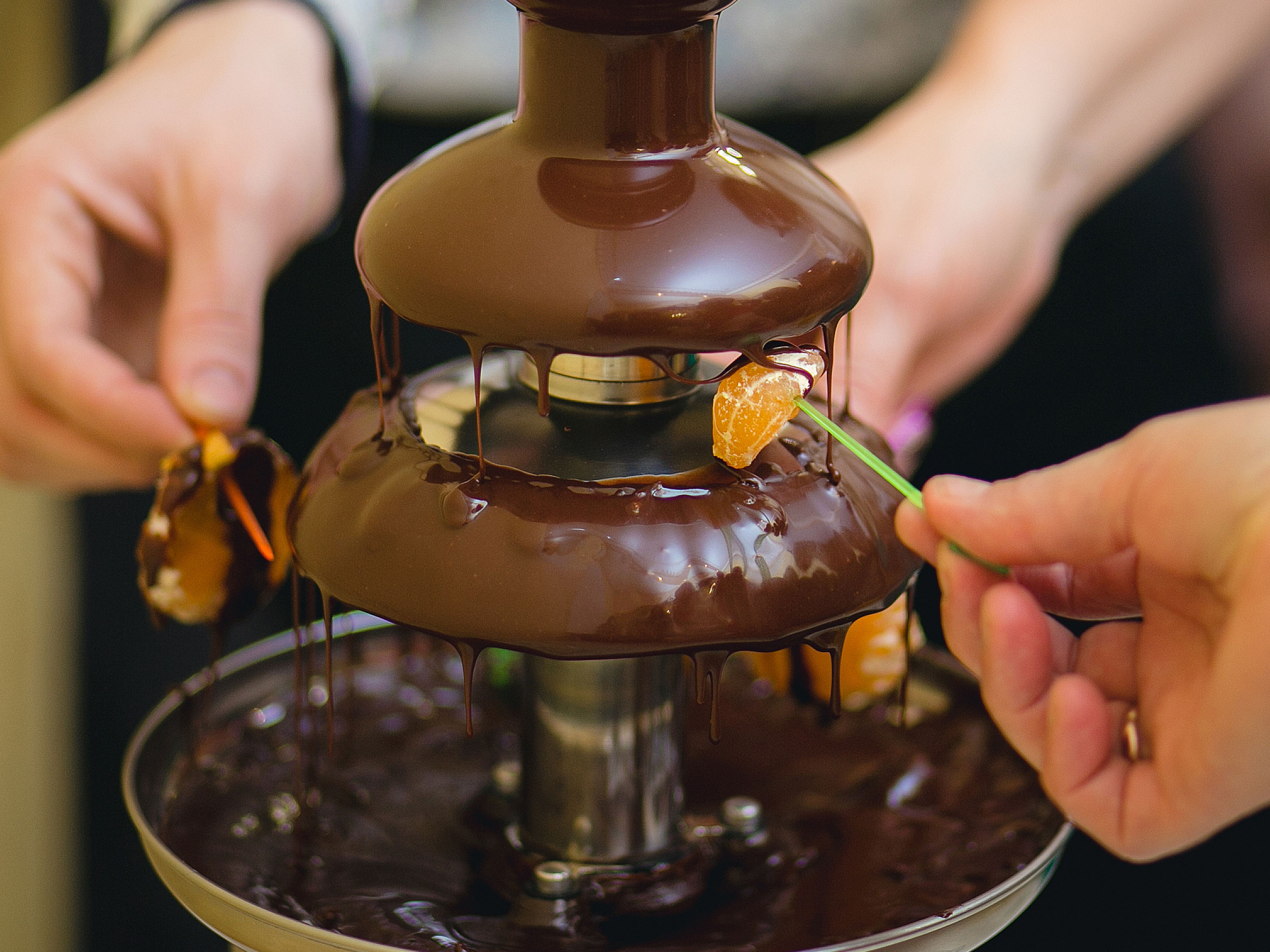 Mani che tengono spiedini di frutta che si immergono in una fontana di cioccolato. Cioccolato fondente scorre giù dalla fontana.
