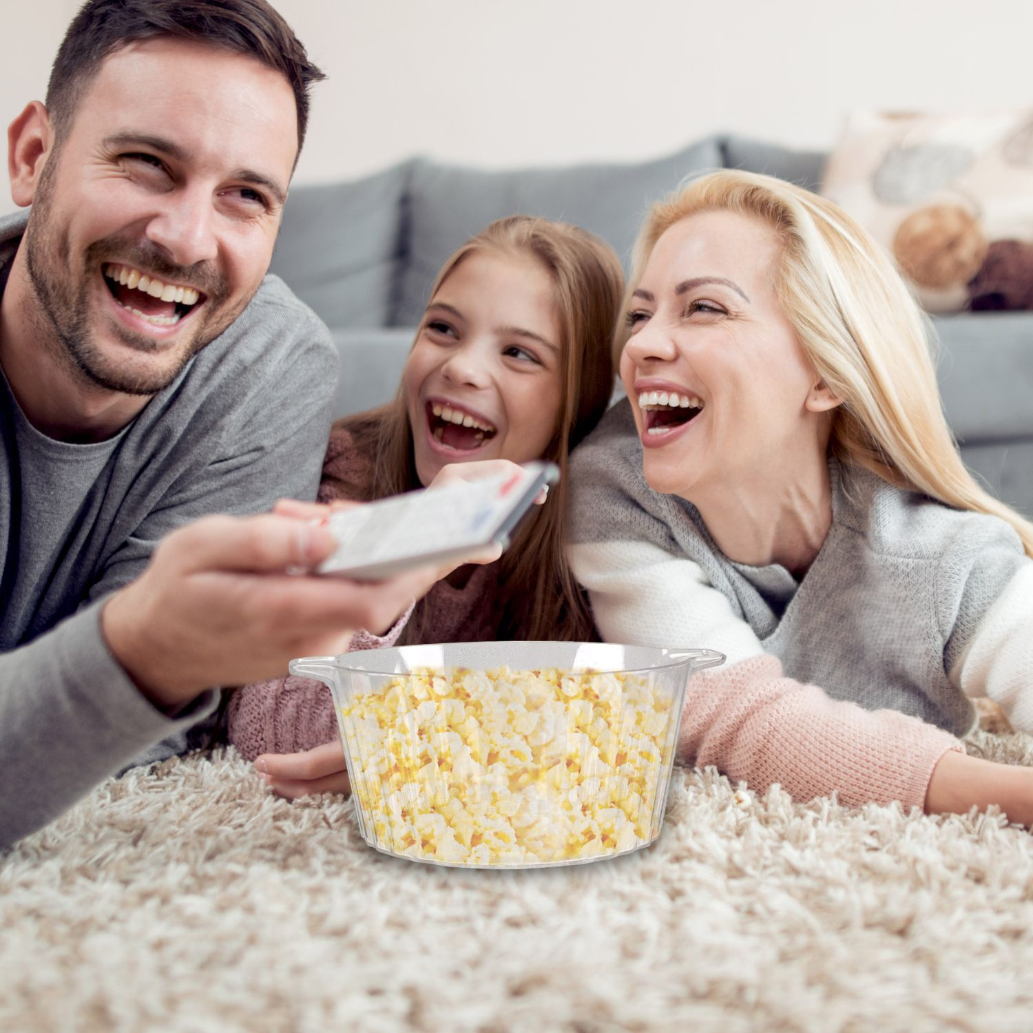 La famiglia ride guardando la TV. Una ciotola di popcorn sul tappeto. Il padre tiene un telecomando.