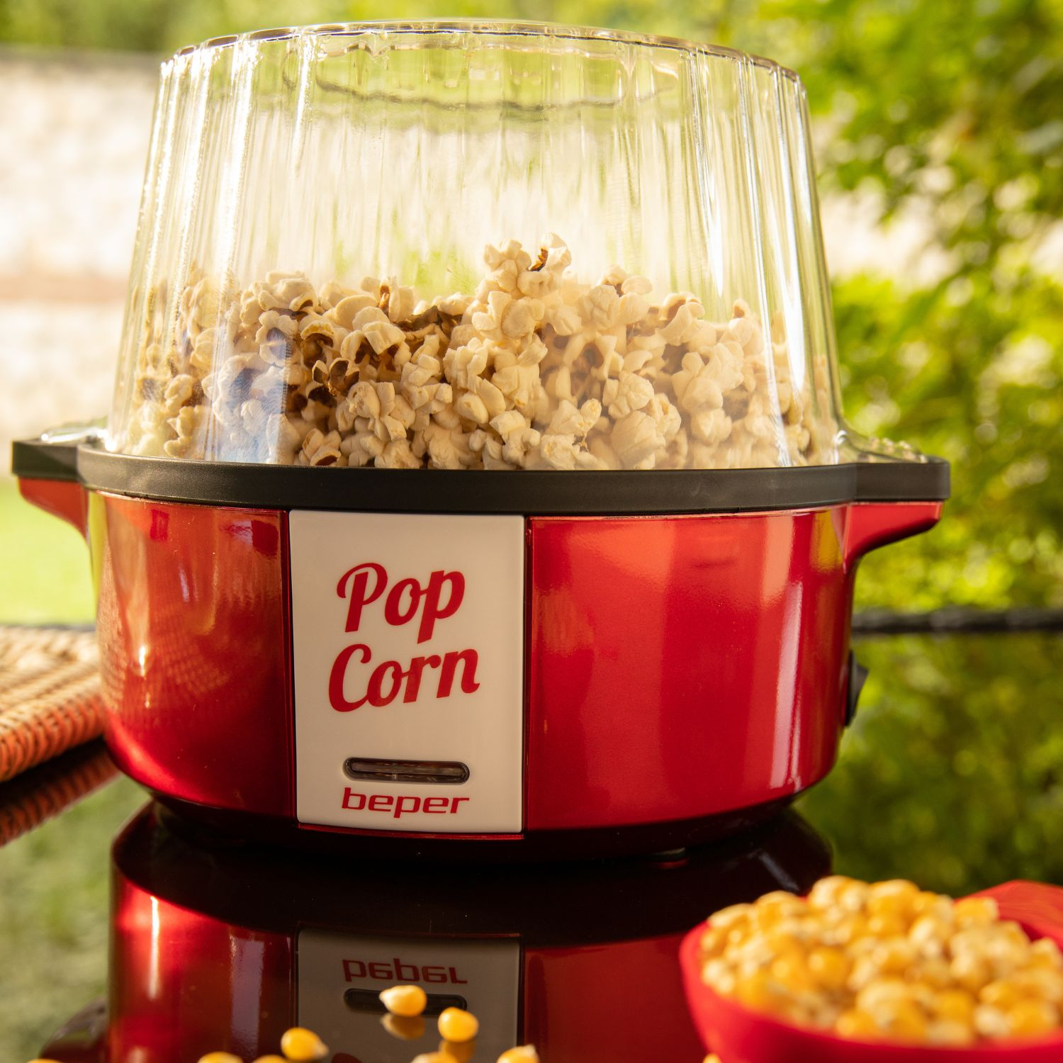 Una macchina per popcorn rossa piena di popcorn, con coperchio trasparente. Lo sfondo mostra fogliame verde e chicchi di mais.