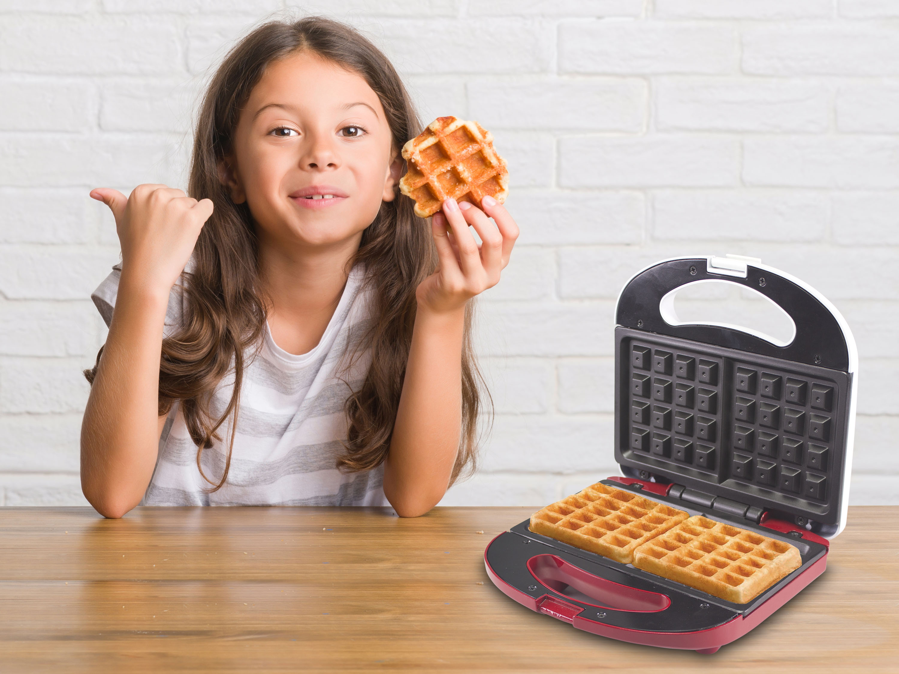 Una ragazza tiene in mano un waffle, indicando un'apparecchiatura per waffle con waffle all'interno. Sfondo: muro di mattoni.