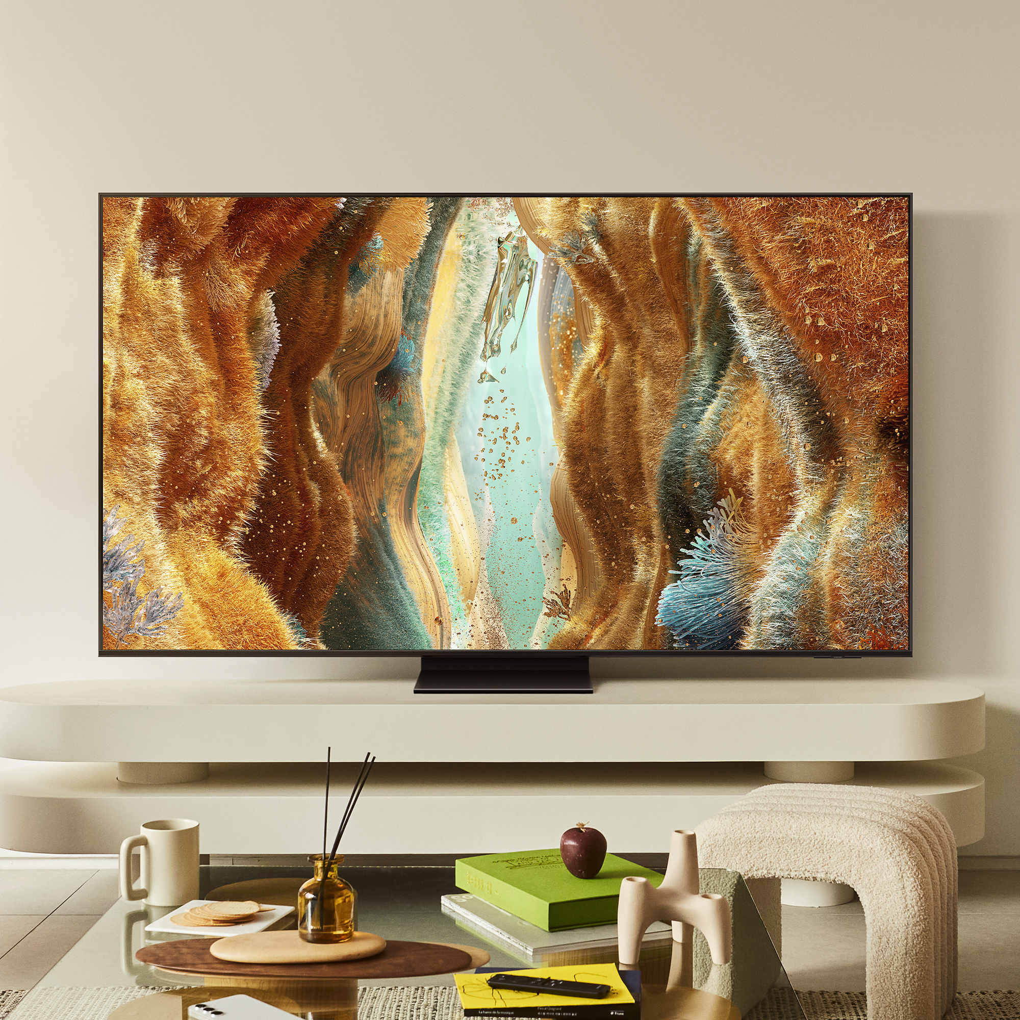 Un grande televisore mostra un'opera d'arte astratta. Un tavolino ha libri, una tazza e un oggetto decorativo. Sfondo beige.
