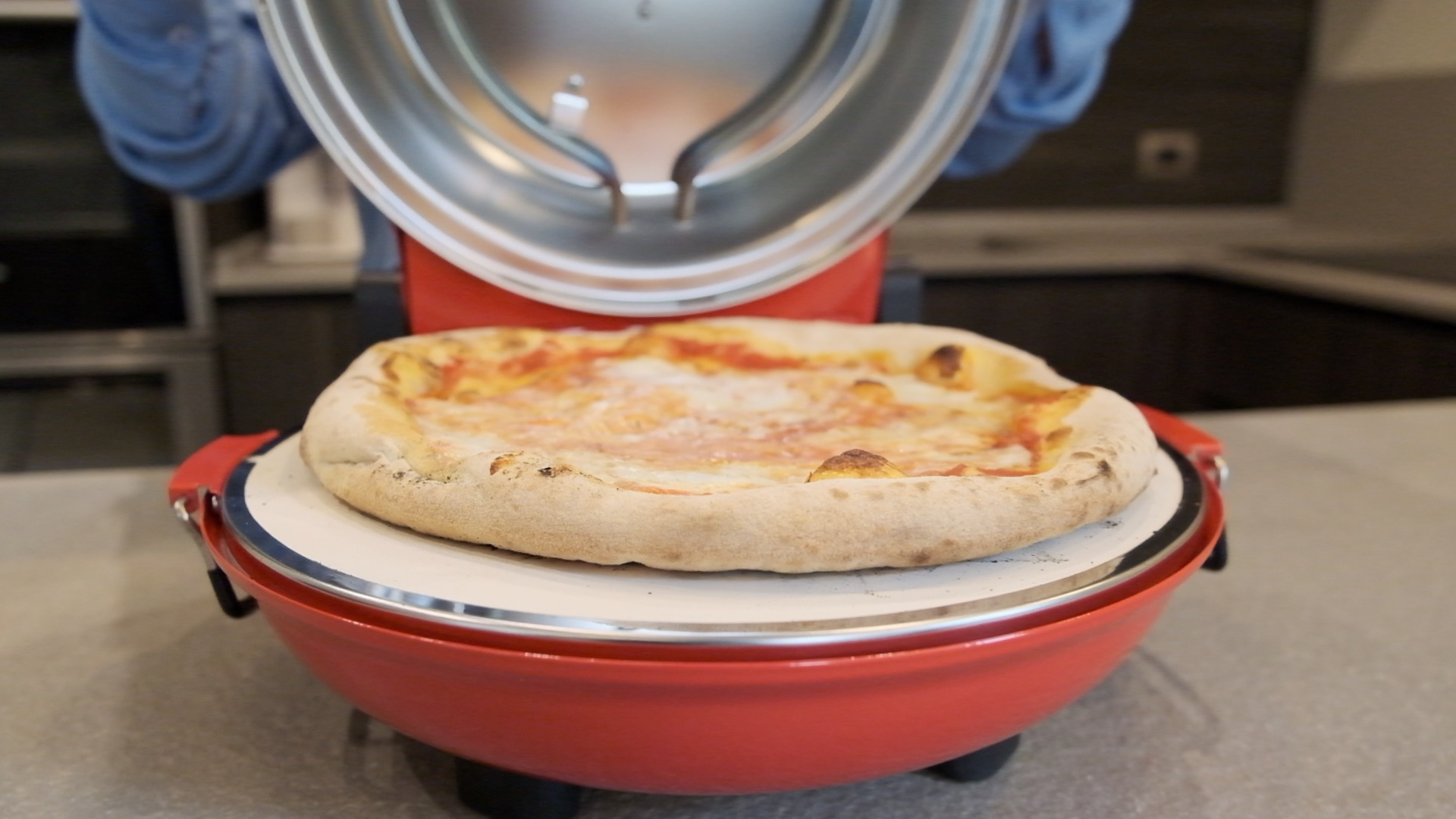 Una pizza cotta in un forno per pizza rosso. Una persona sta aprendo il forno. Lo sfondo è una cucina.