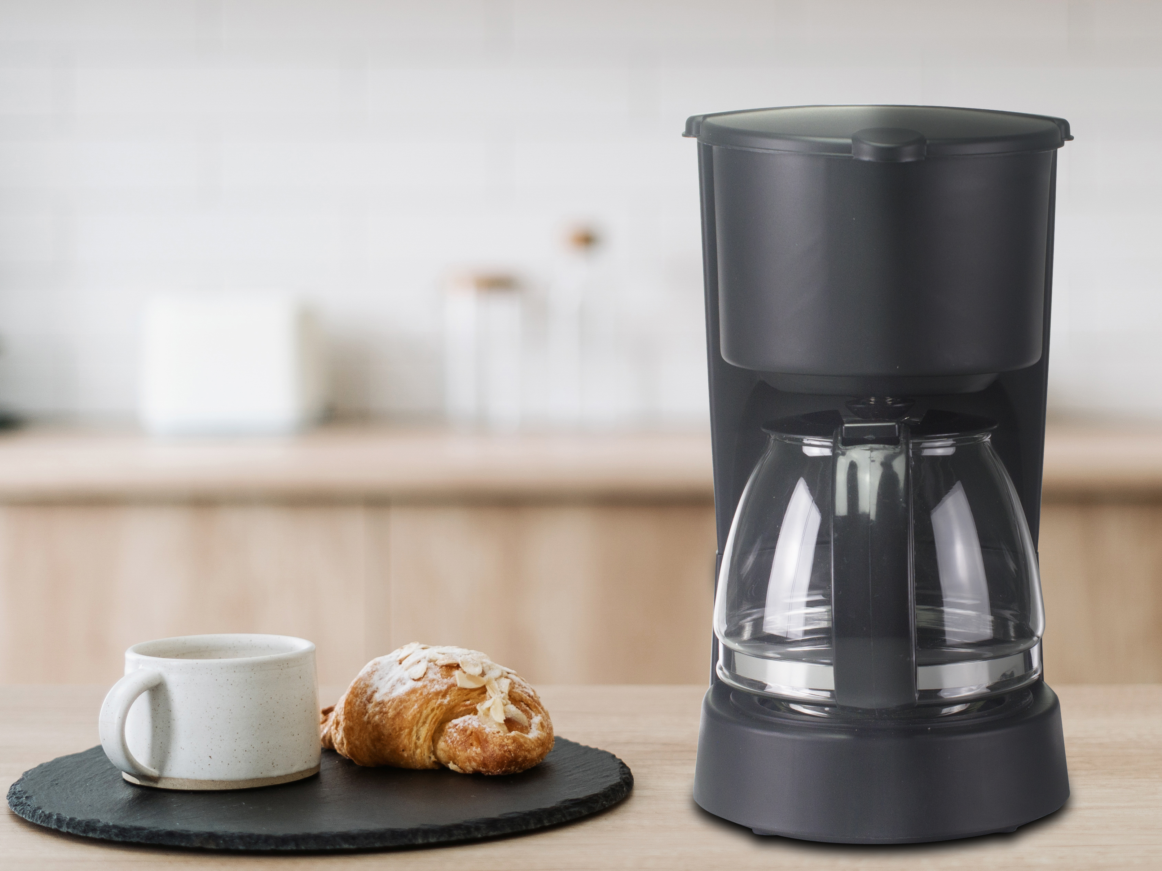Una caffettiera nera, un croissant e una tazza sono su un tavolo di legno. Lo sfondo mostra una cucina.