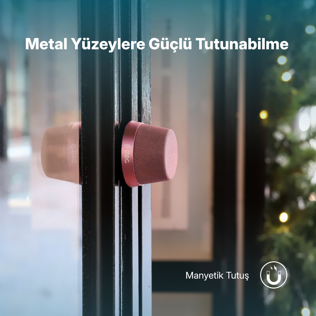 Siyah metal yüzeye bağlı pembe bir hoparlör. Metin 'Metal Yüzeylere Yapışma' diyor. Arka plan bulanık.