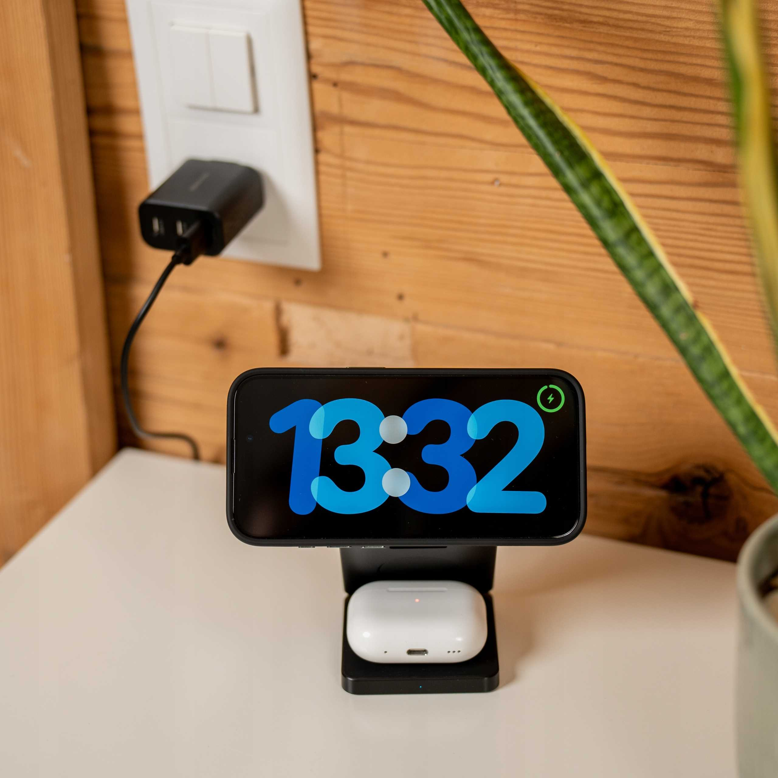 Telefon wyświetlający 13:32 na stacji ładującej z AirPods. Gniazdko ścienne i roślina w tle.