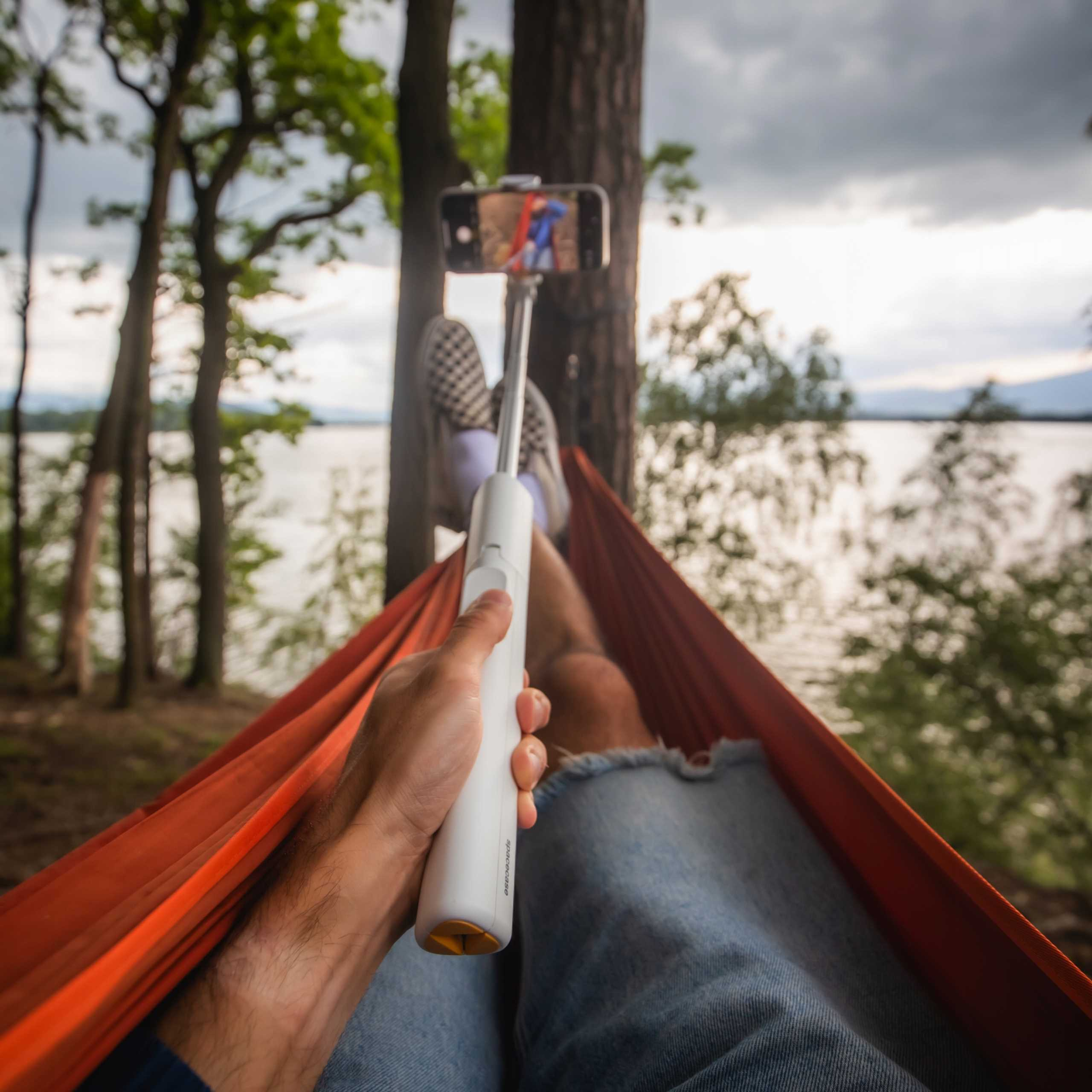 Osoba w hamaku robi selfie telefonem na patyku do selfie. Jezioro i drzewa w tle.