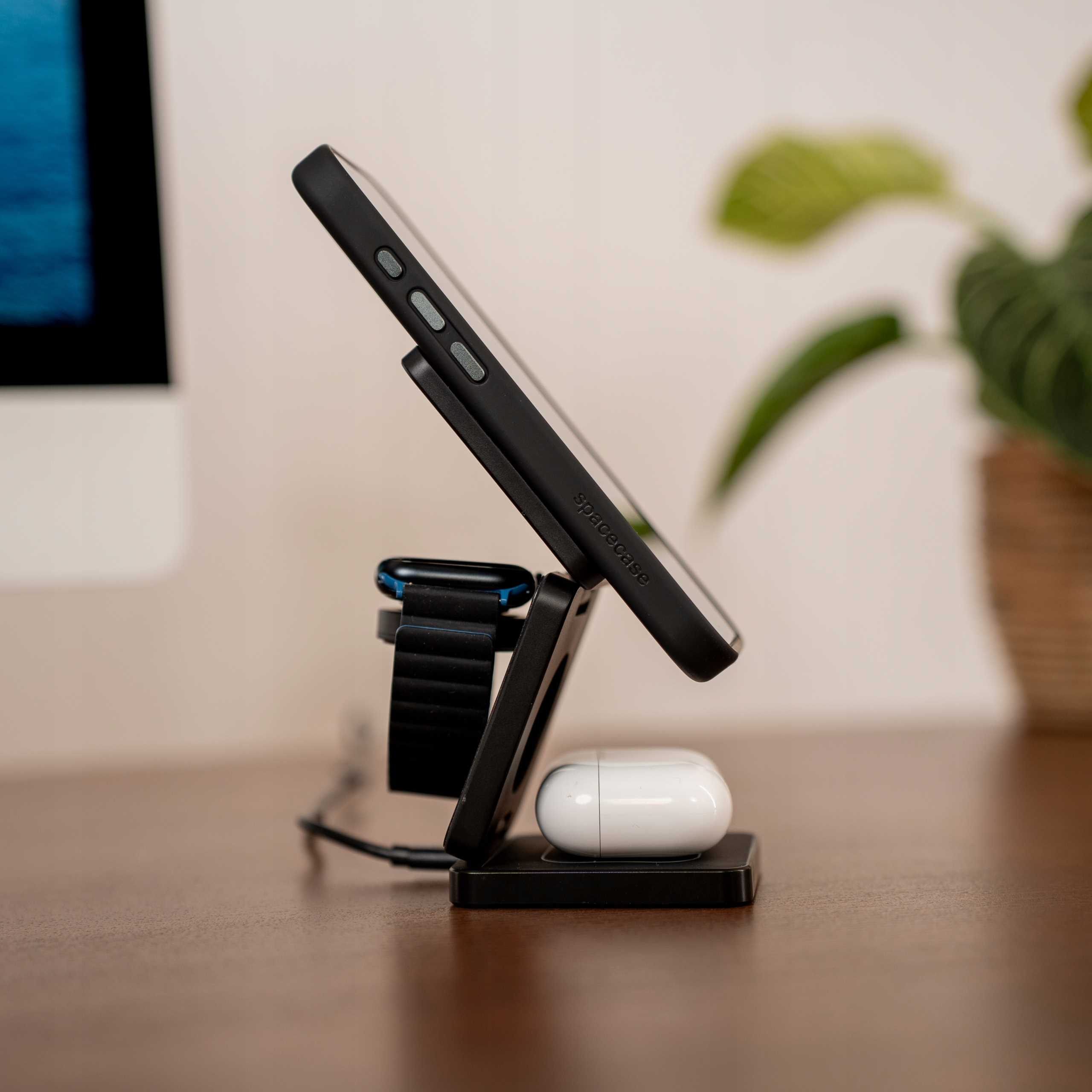 Czarny stojak ładujący z telefonem, zegarkiem i słuchawkami. Roślina i komputer w tle.