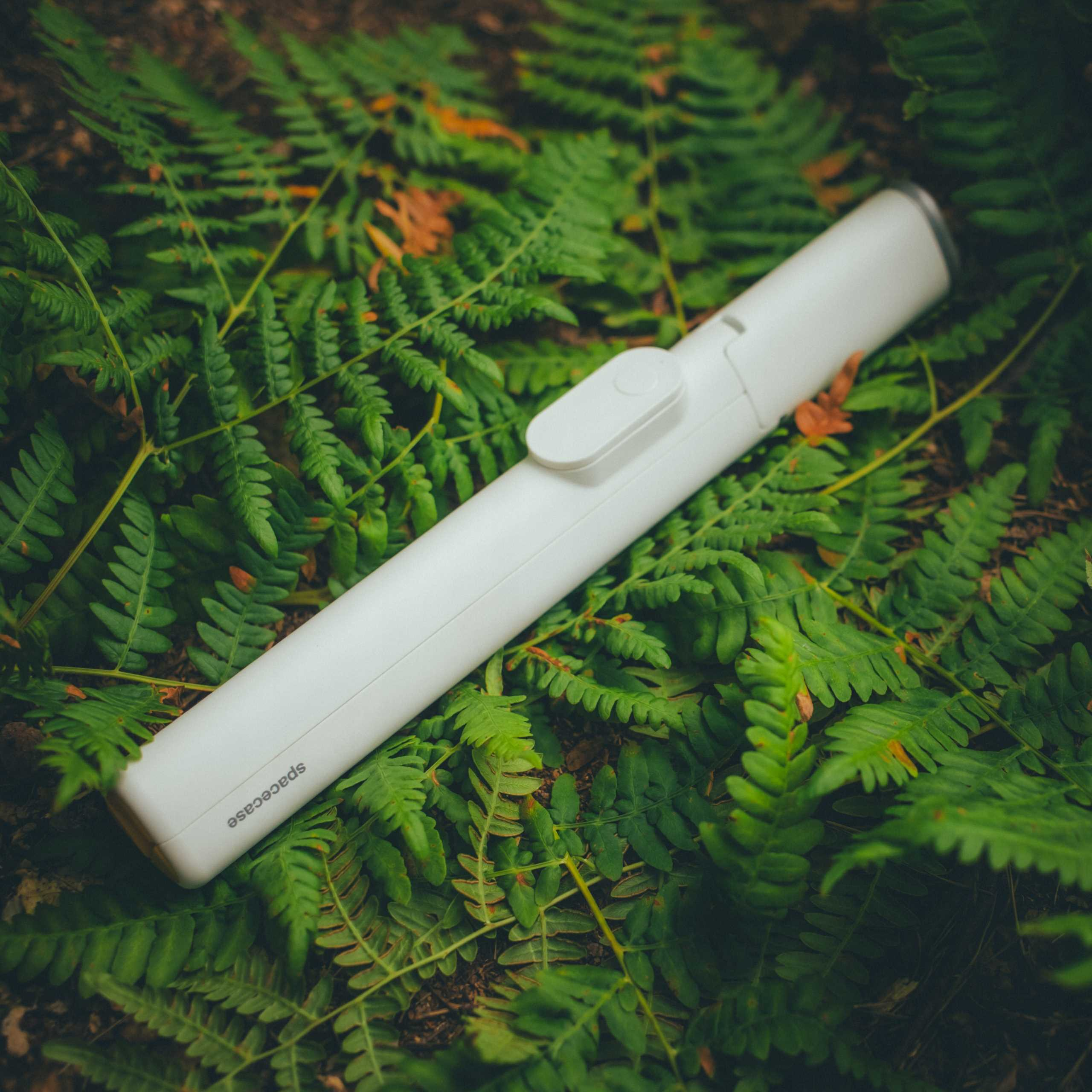 Biały gadżet Spacecase na zielonych paprociach. Gadżet ma przycisk i nazwę marki.