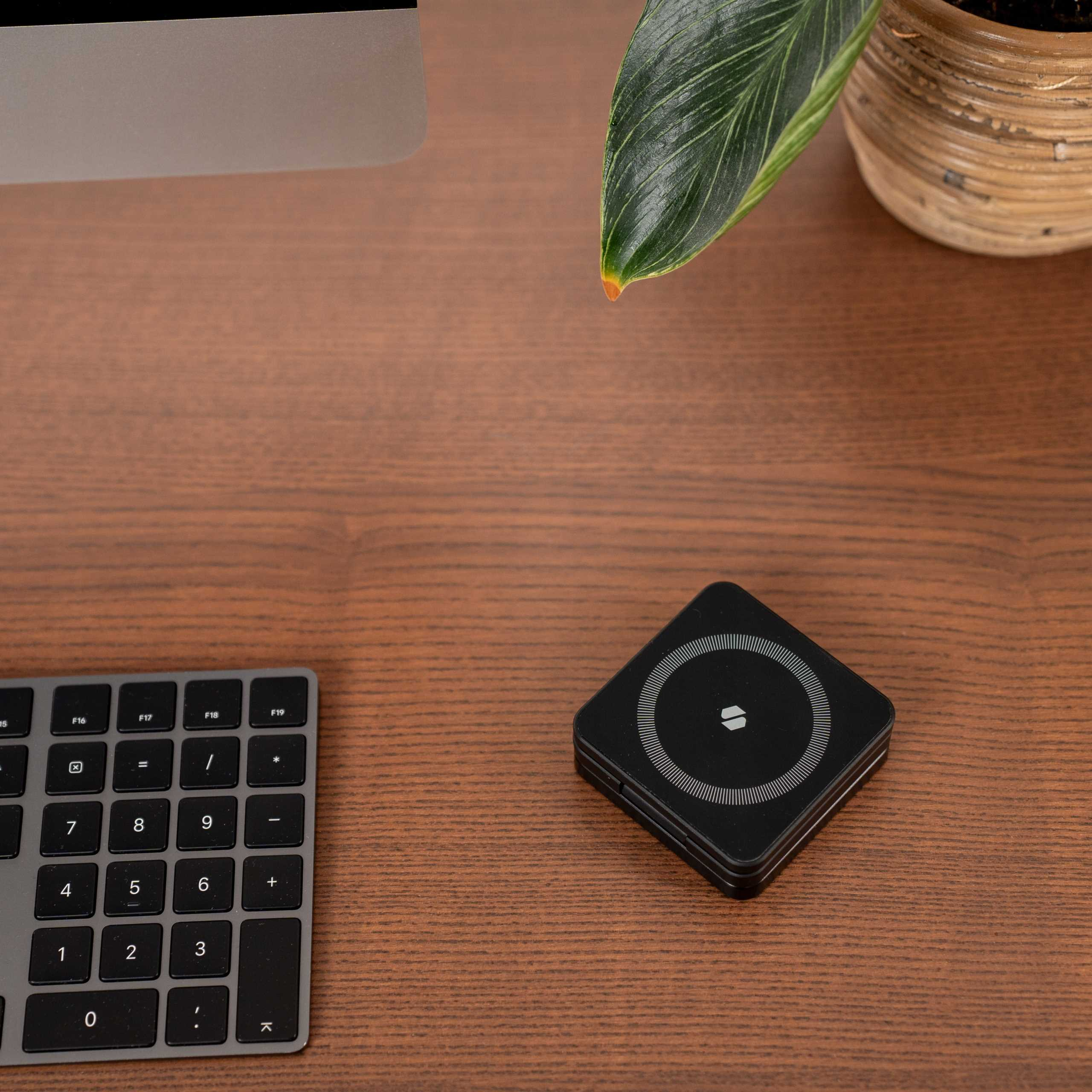 Drewniane biurko z klawiaturą, czarnym urządzeniem i rośliną w doniczce. Ekran komputera jest w tle.