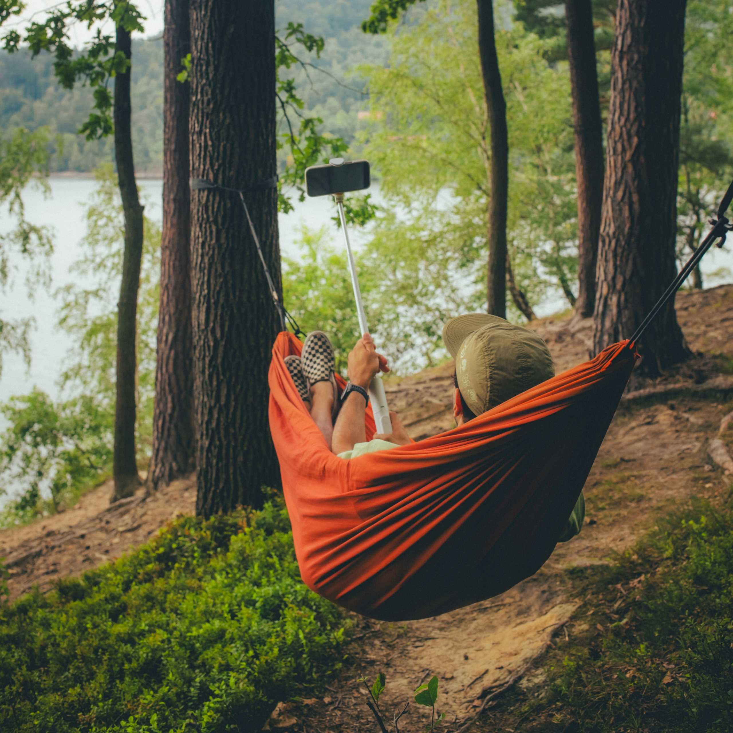 Osoba w pomarańczowym hamaku, trzymająca kij do selfie z telefonem. Tło lasu, widoczne drzewa, woda i zieleń.