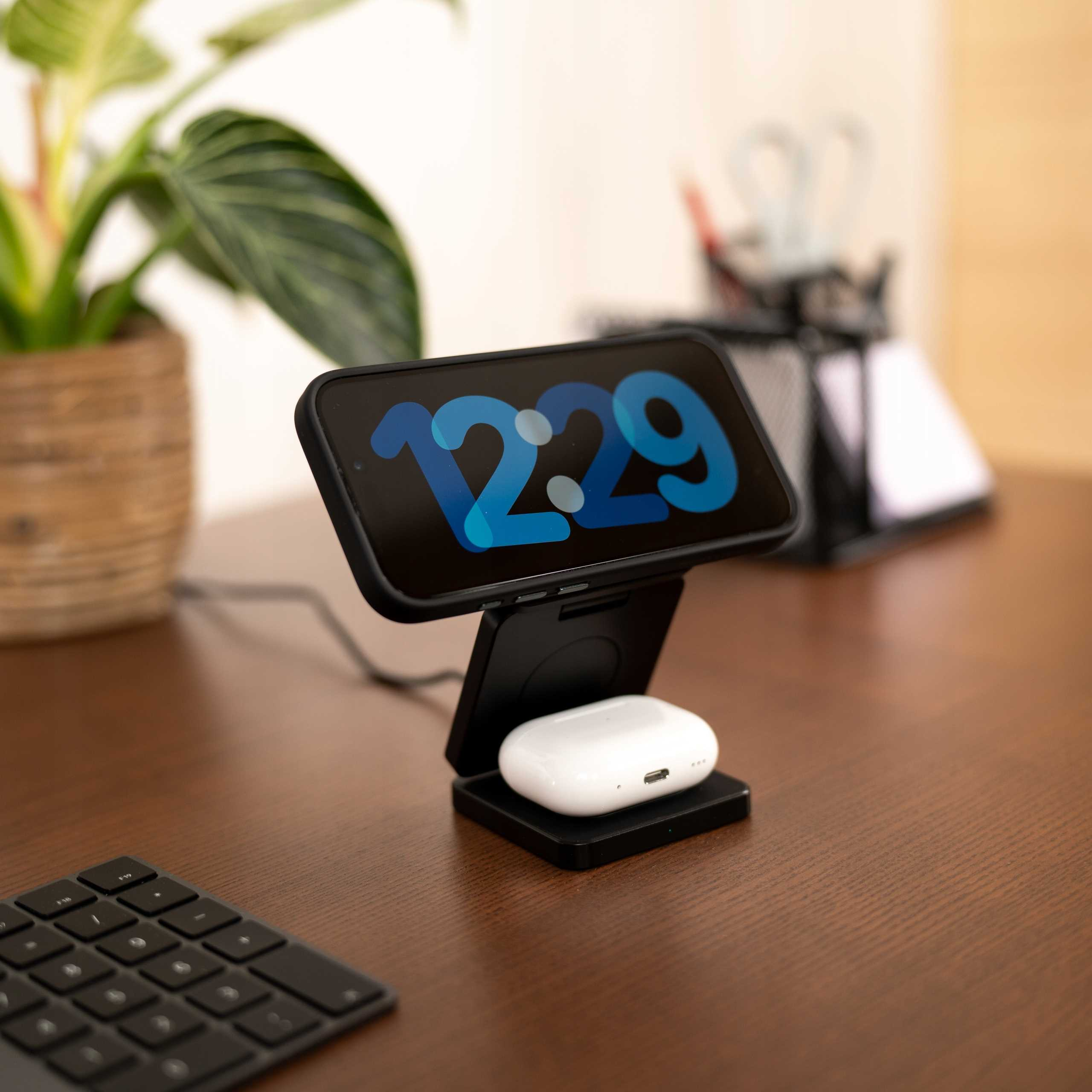 Telefon z godziną 12:29 spoczywa na czarnej podstawce ładującej. Białe słuchawki poniżej. Roślina i klawiatura w zasięgu wzroku.