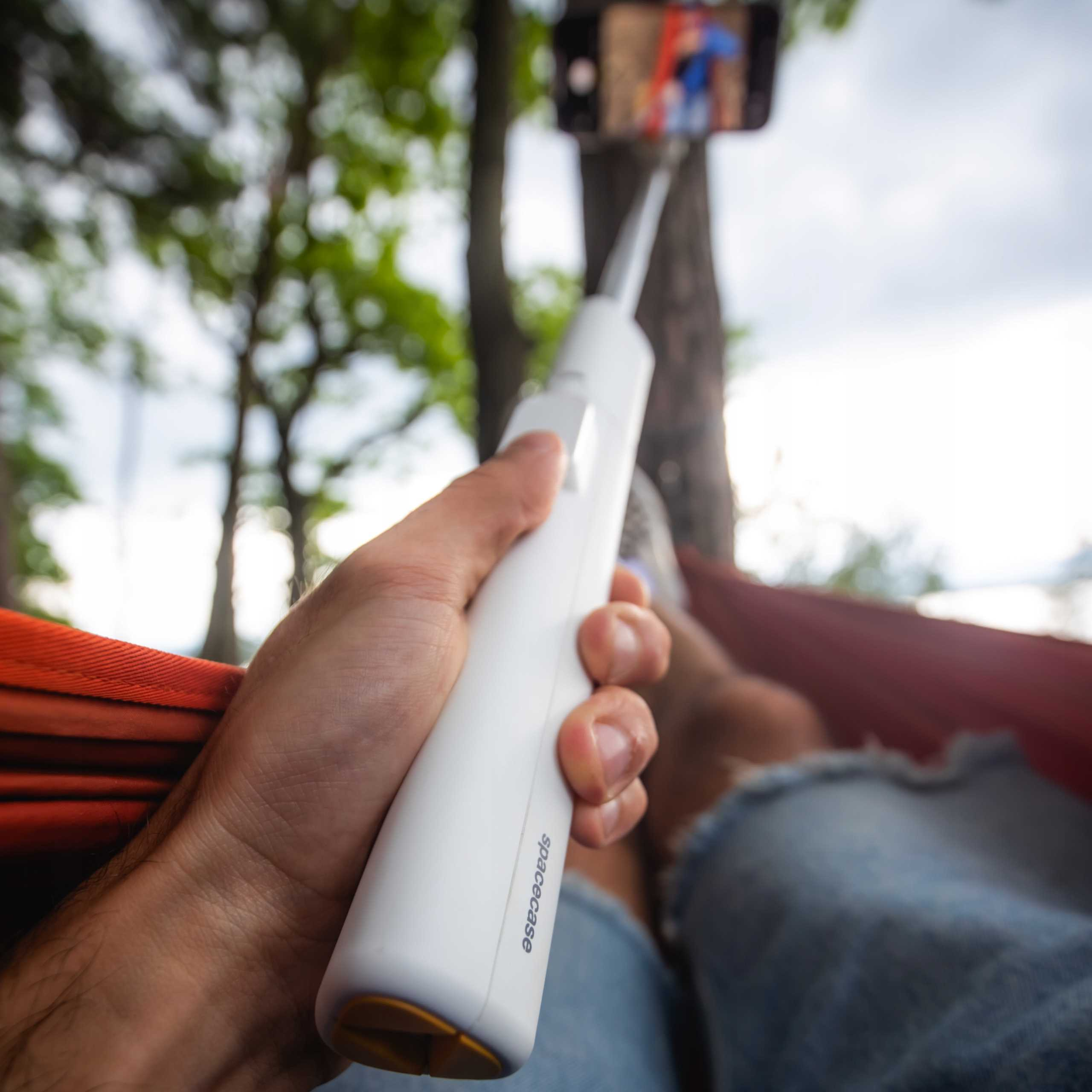 Osoba trzyma biały kijek do selfie z dołączonym telefonem. Relaksują się w hamaku w lesie.