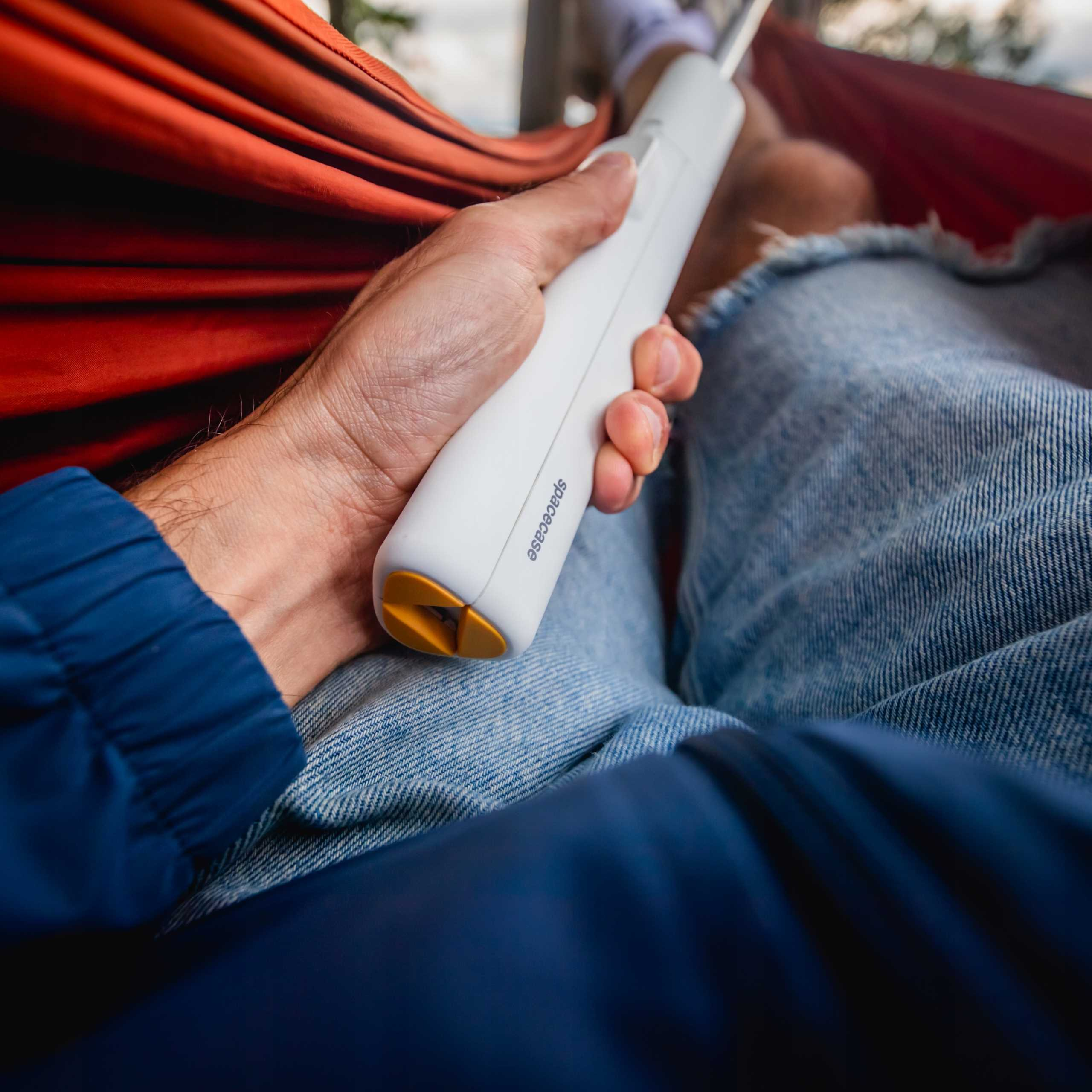 Osoba trzyma białe urządzenie w hamaku. Widoczne są dżinsy, niebieska kurtka i czerwony hamak.