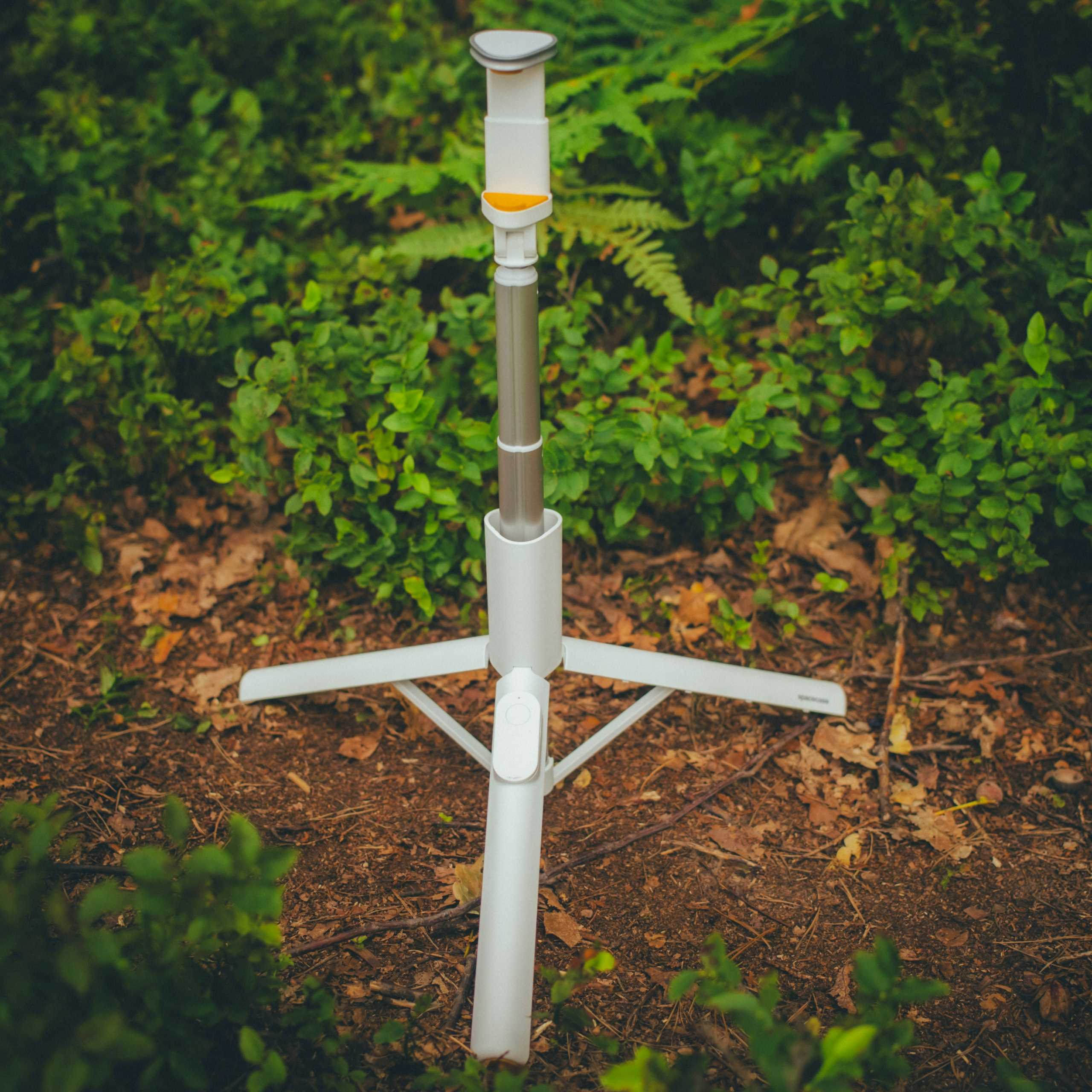 Biały statyw stoi na brązowej leśnej podłodze, otoczony zielonymi liśćmi. To styl selfie stick.