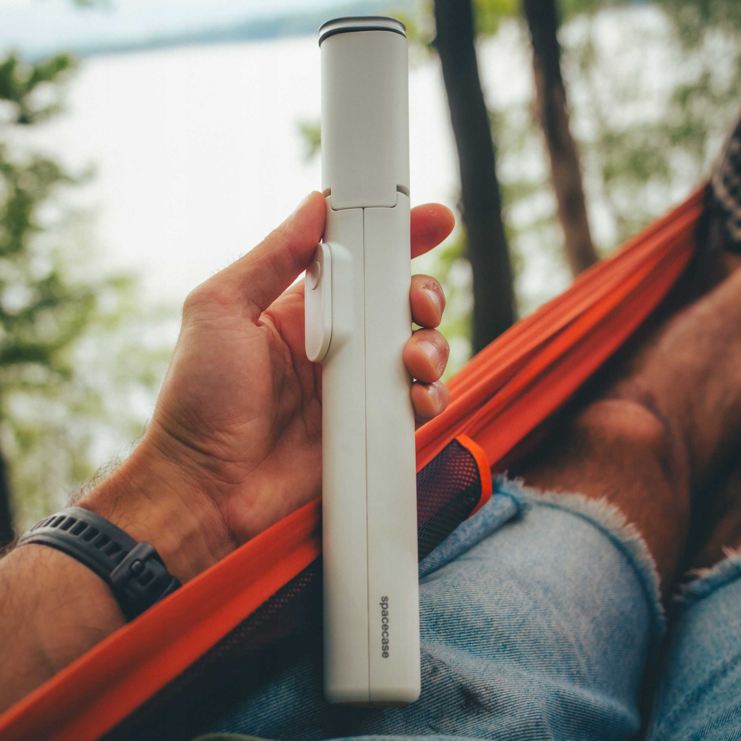 Ręka trzyma białe urządzenie Spacecase. Osoba jest w pomarańczowym hamaku, w dżinsach. W tle drzewa i woda.
