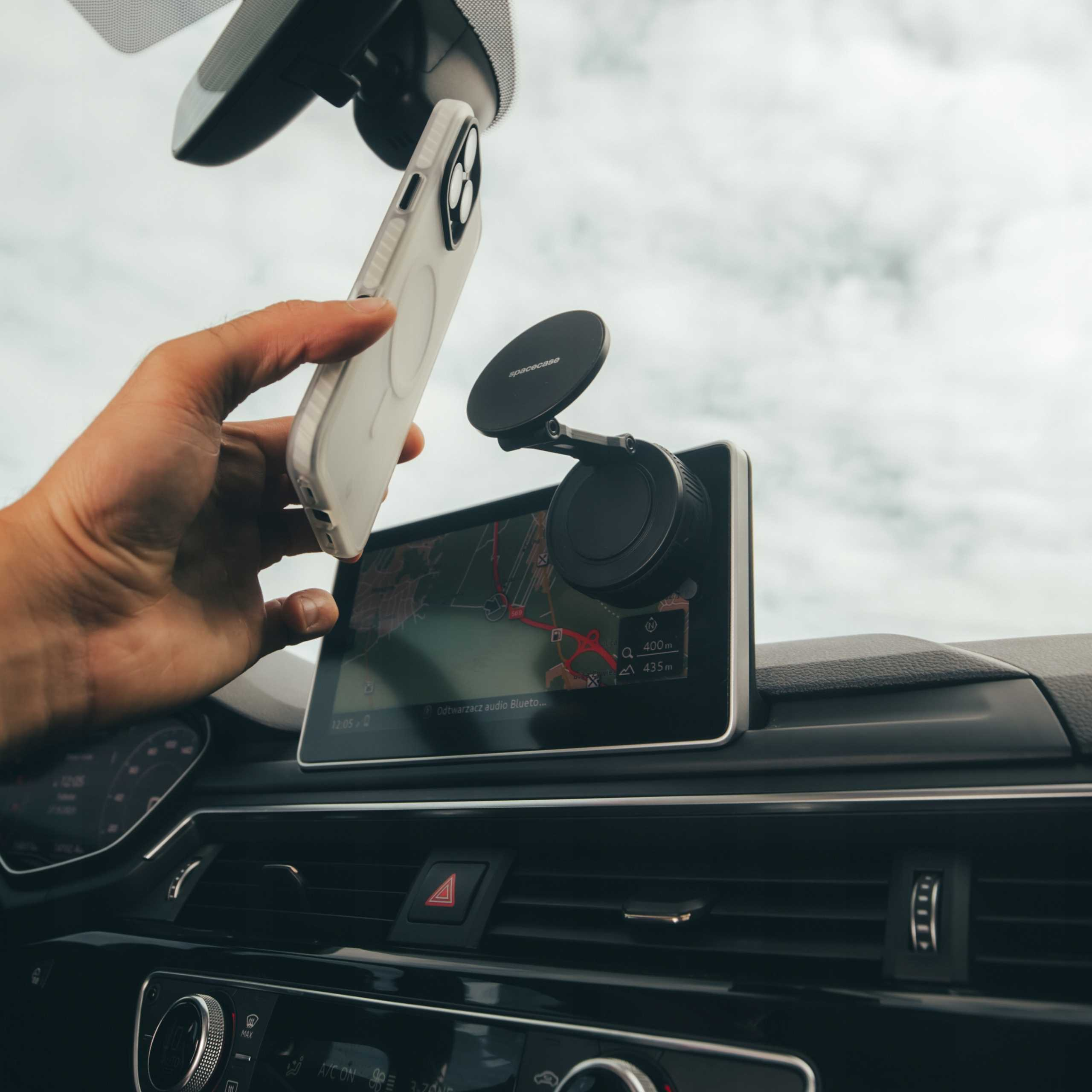 Ręka trzymająca telefon w pobliżu deski rozdzielczej samochodu z ekranem GPS i uchwytem na telefon.