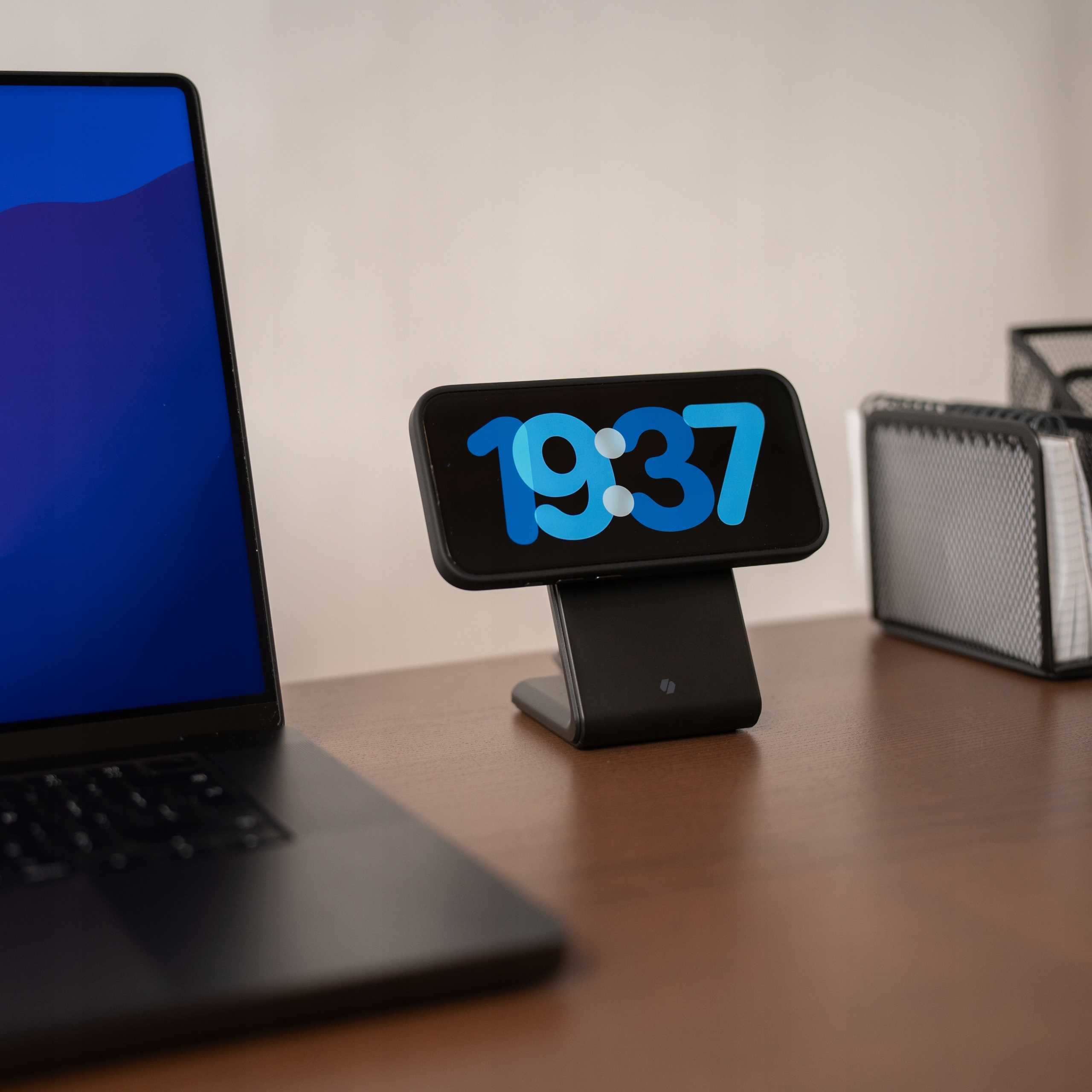 Telefon ładujący się, wyświetlający 19:37, na biurku. Z lewej strony laptop. Z tyłu organizer z siatki drucianej.