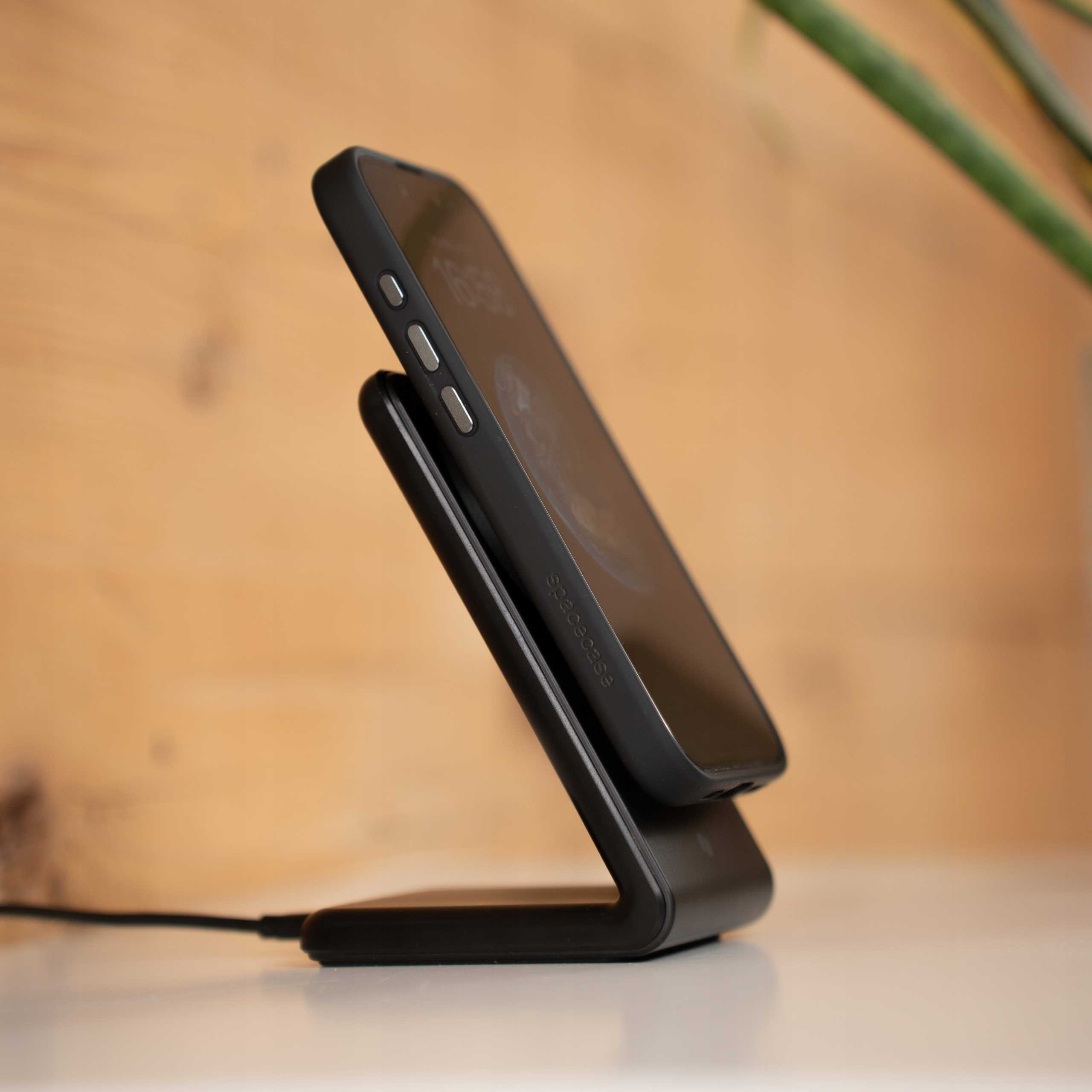 Czarny telefon leży na czarnej ładowarce bezprzewodowej. Tło przedstawia drewnianą ścianę i roślinę.
