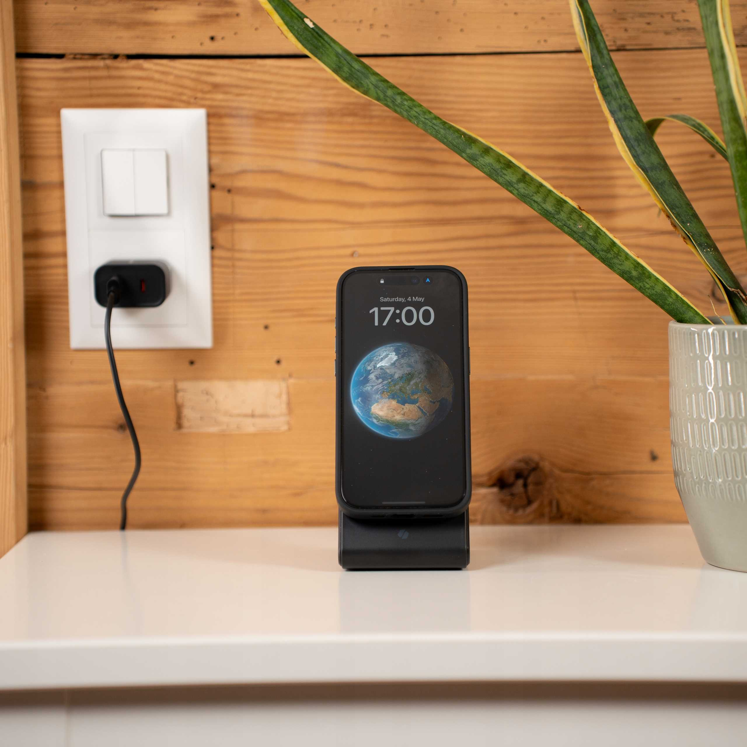 Telefon pokazujący Ziemię, ładujący się na czarnej stacji dokującej. Po prawej roślina. Ładowarka jest podłączona do ściany.