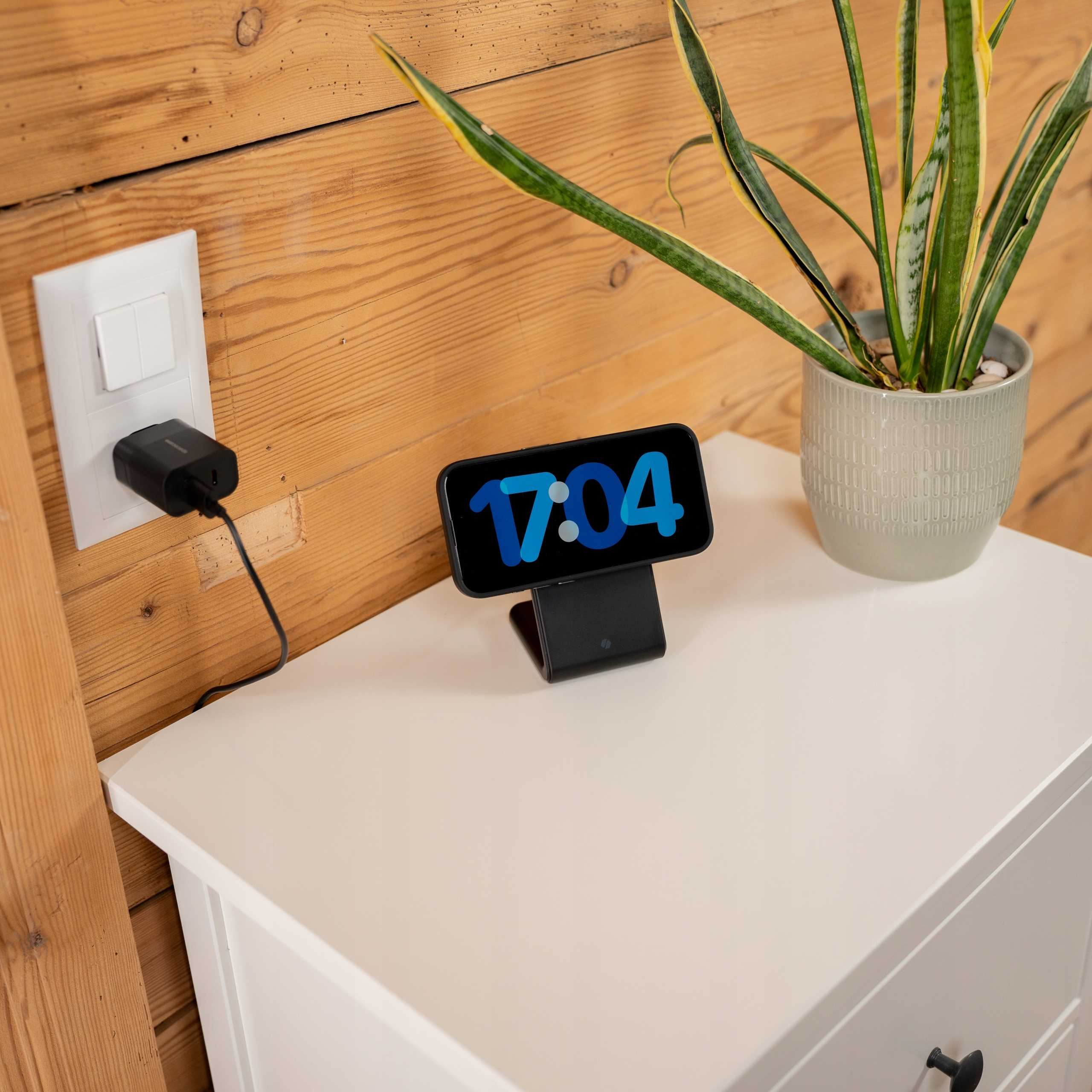 Telefon na ładowarce wyświetla godzinę 17:04. Obok niego stoi roślina na białym stole. Gniazdko elektryczne jest na ścianie.